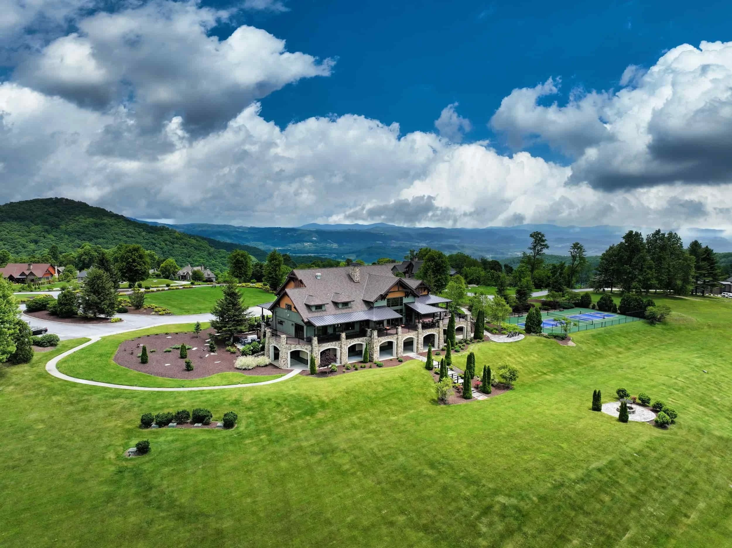 Mountain Lodge in Grand Highlands at Bearwallow Mountain Hendersonville NC with stone and wood exterior, surrounded by landscaped lawn and gardens, tennis courts, trees, and rolling hills with mountains in the background under partly cloudy skies.