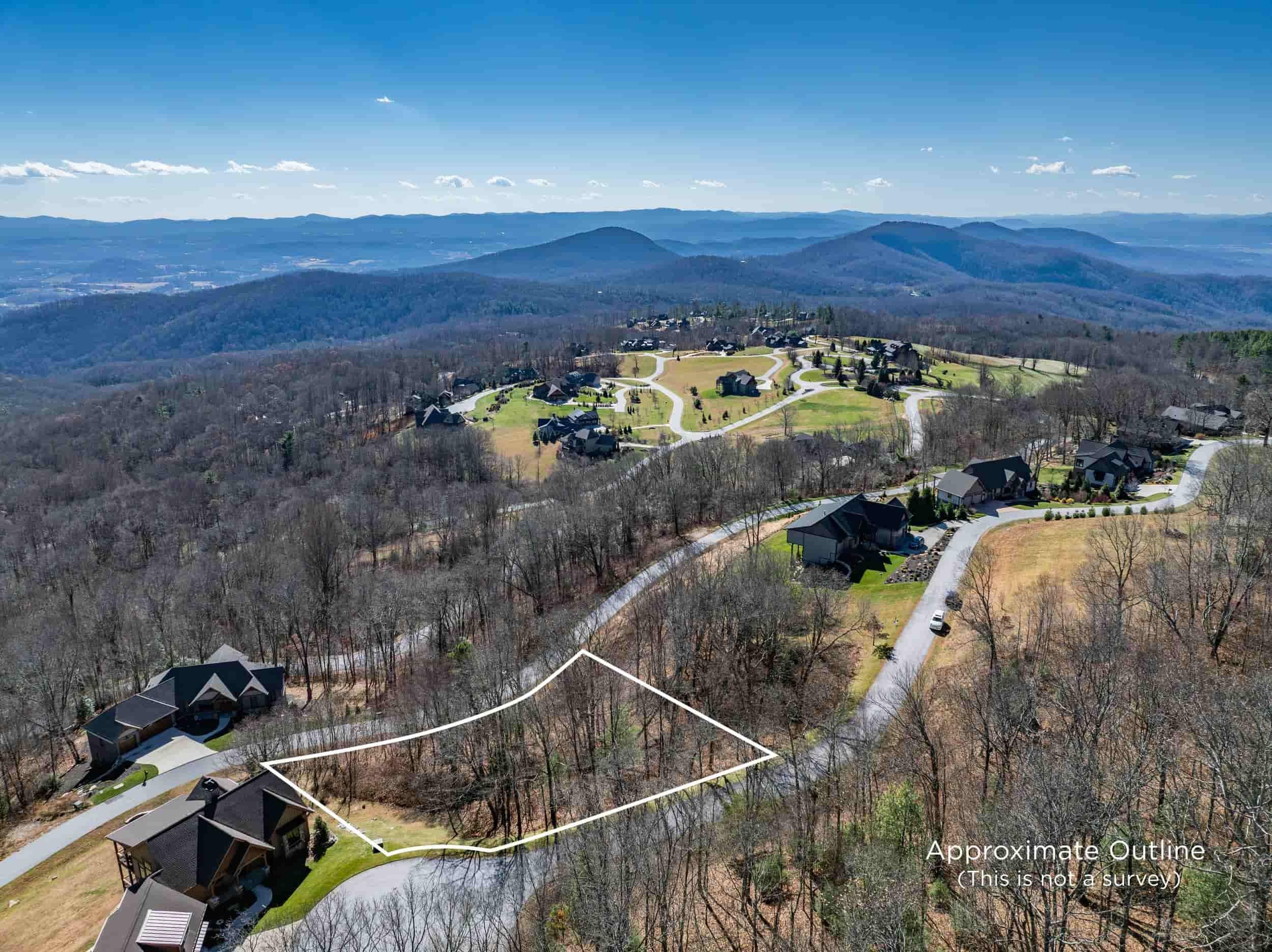Aerial view of a lot for sale with Ashton Peters Realtor in Grand Highlands at Bearwallow Mountain Hendersonville NC with winding roads and houses surrounded by trees, with distant mountains in the background under a blue sky.