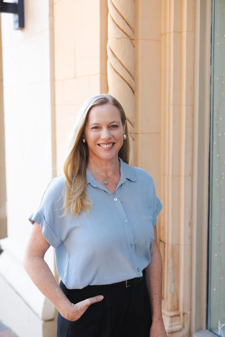Lauren Butcher Realtor wearing a light blue short-sleeved blouse and black pants, standing outside near a building with beige stone details, smiling at the camera.