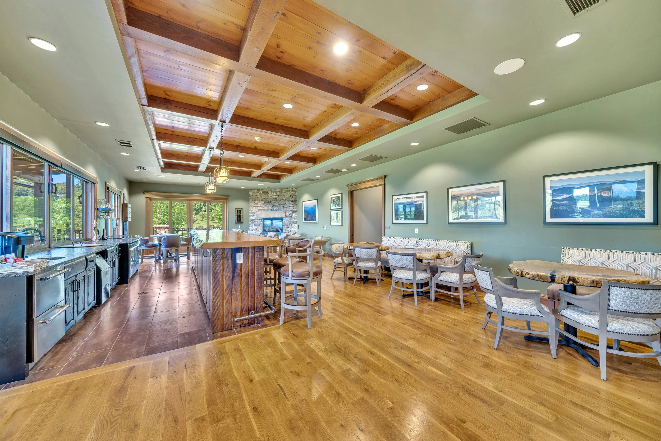 Interior of a spacious room with a wooden ceiling, large windows, and a wood-paneled bar area with chairs. There are tables and chairs along the green wall with framed pictures, and a stone fireplace at the back.