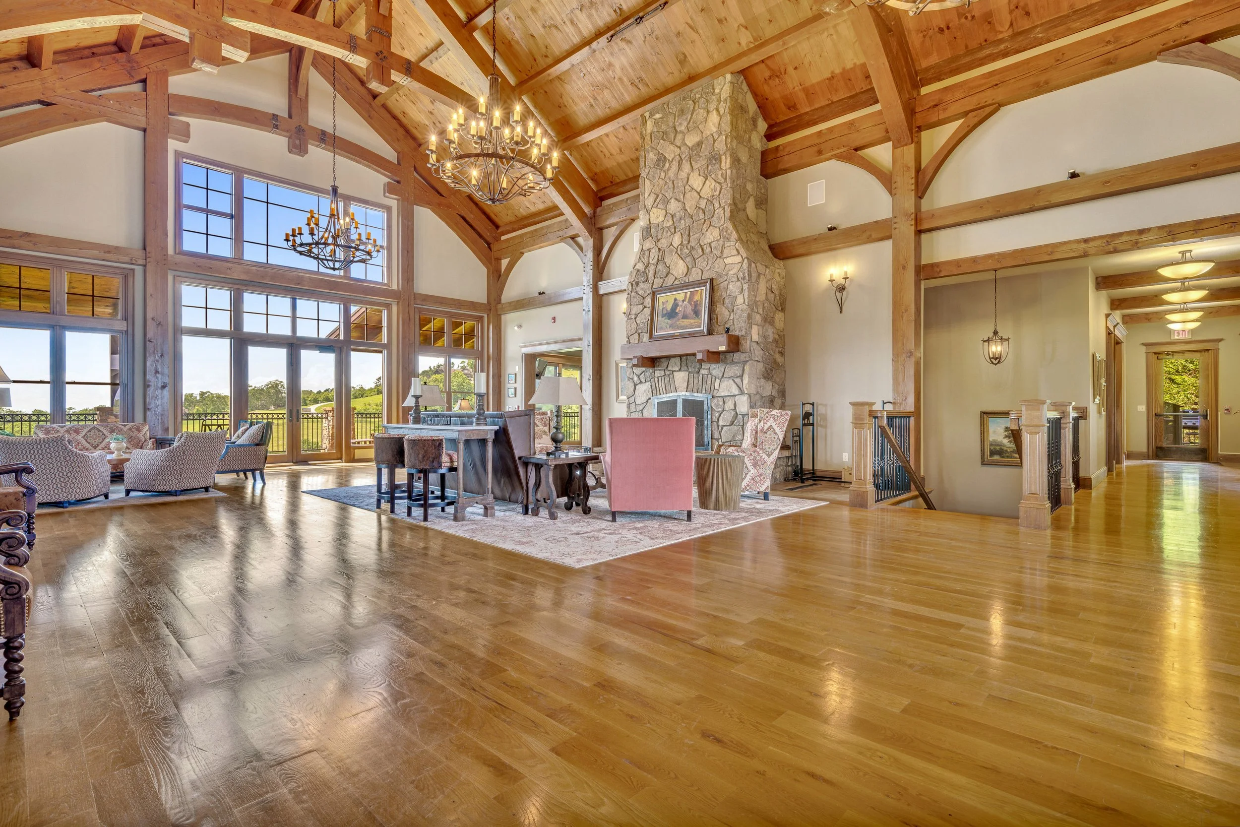 Spacious living room with hardwood floors, large windows, stone fireplace, high wooden ceiling, chandeliers, and seating area.
