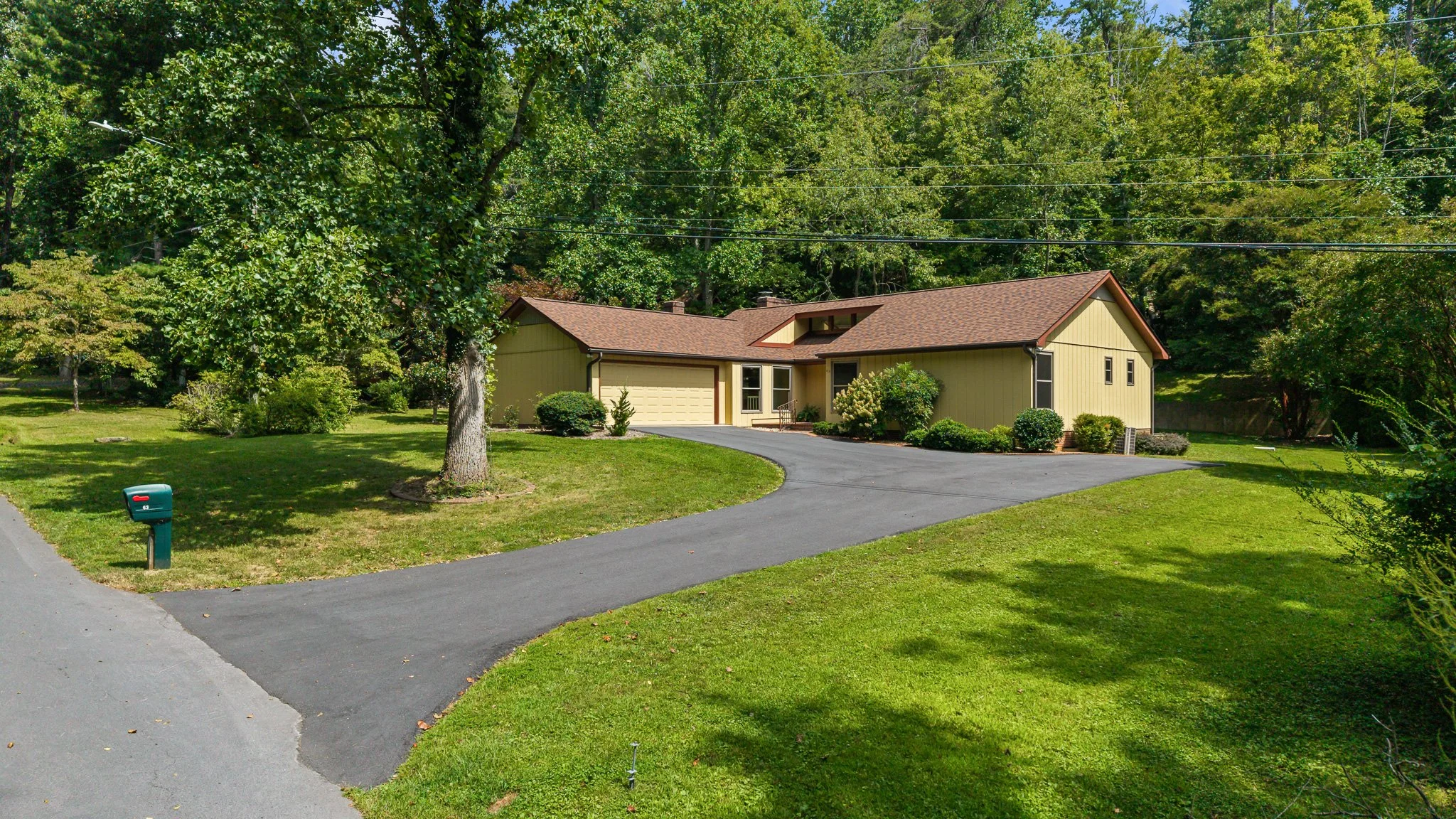 A single-story yellow home for sale with Ashton Peters Realtor in Asheville NC with a brown roof, surrounded by a well-maintained lawn and shrubs, with a wooded background.