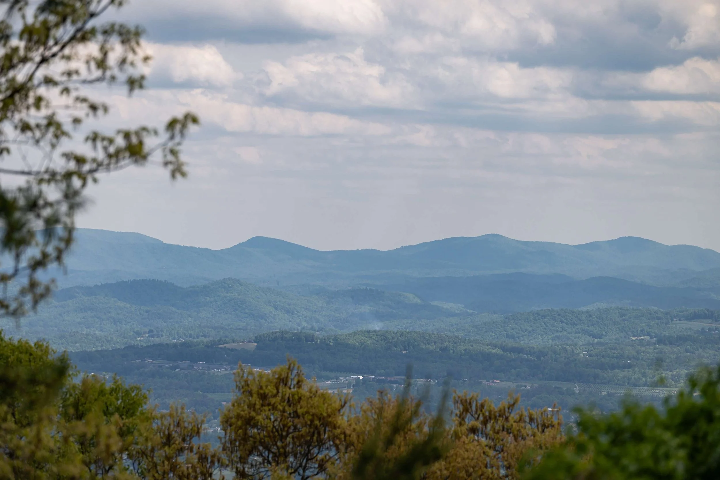 Aerial view of a lot for sale with Ashton Peters Realtor in Grand Highlands at Bearwallow Mountain Hendersonville NC with winding roads and houses surrounded by trees, with distant mountains in the background under a blue sky.