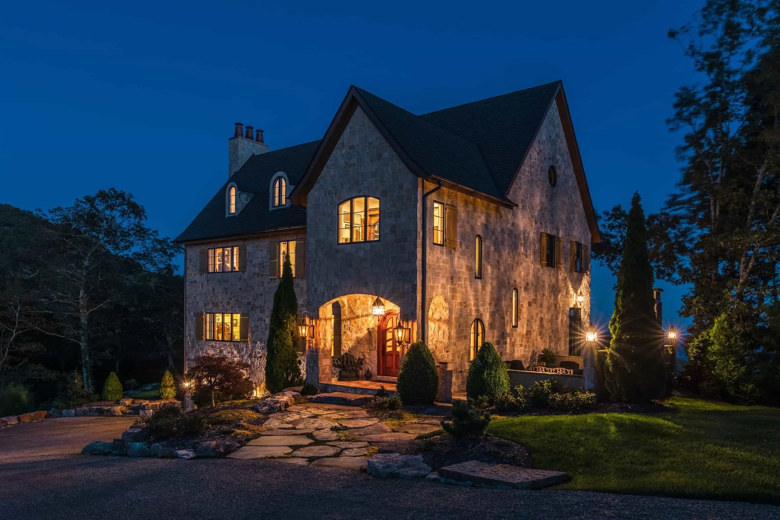 A luxury mountain home in Grand Highlands at Bearwallow Mountain Hendersonville NC sold by Top Listing Agent Ashton Peters Realtor at dusk, with windows and outdoor lights, surrounded by beautiful landscaping.