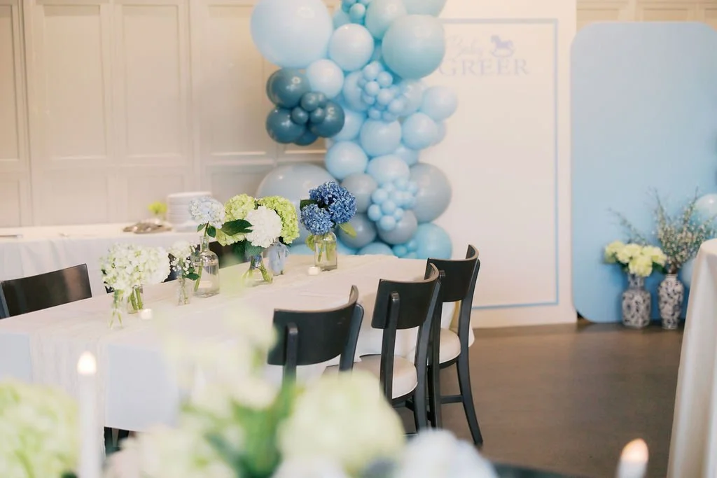 A decorated event room with a table set with small flower arrangements and a balloon display in shades of blue in the background.