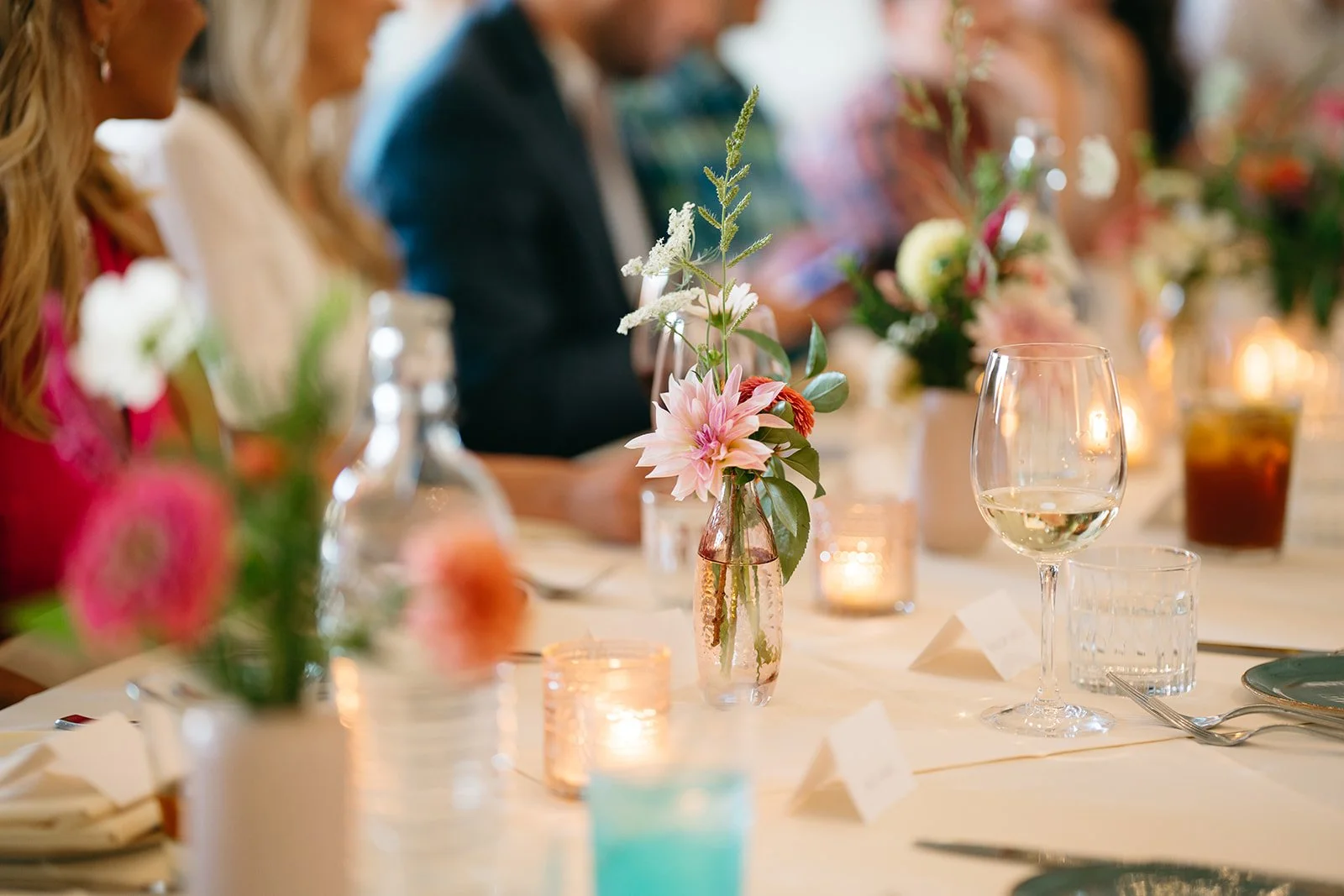 A beautifully arranged table at a formal event with pink and white flowers in vases, candles, wine glasses, and neatly folded napkins, with guests seated in the background.