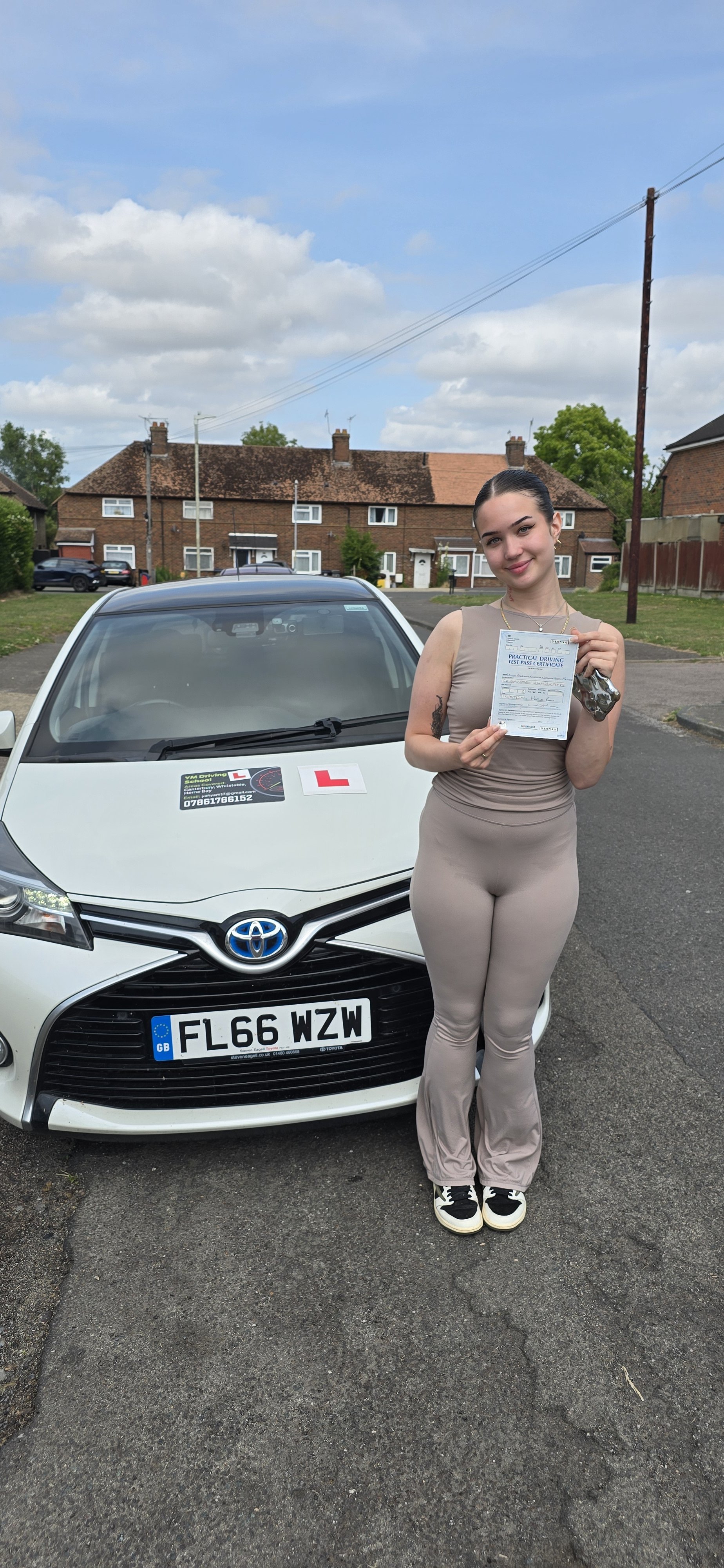Young woman holding a driving test certificate standing next to a white Toyota car with a learner's permit sign, on a residential street with houses and a cloudy sky in the background.