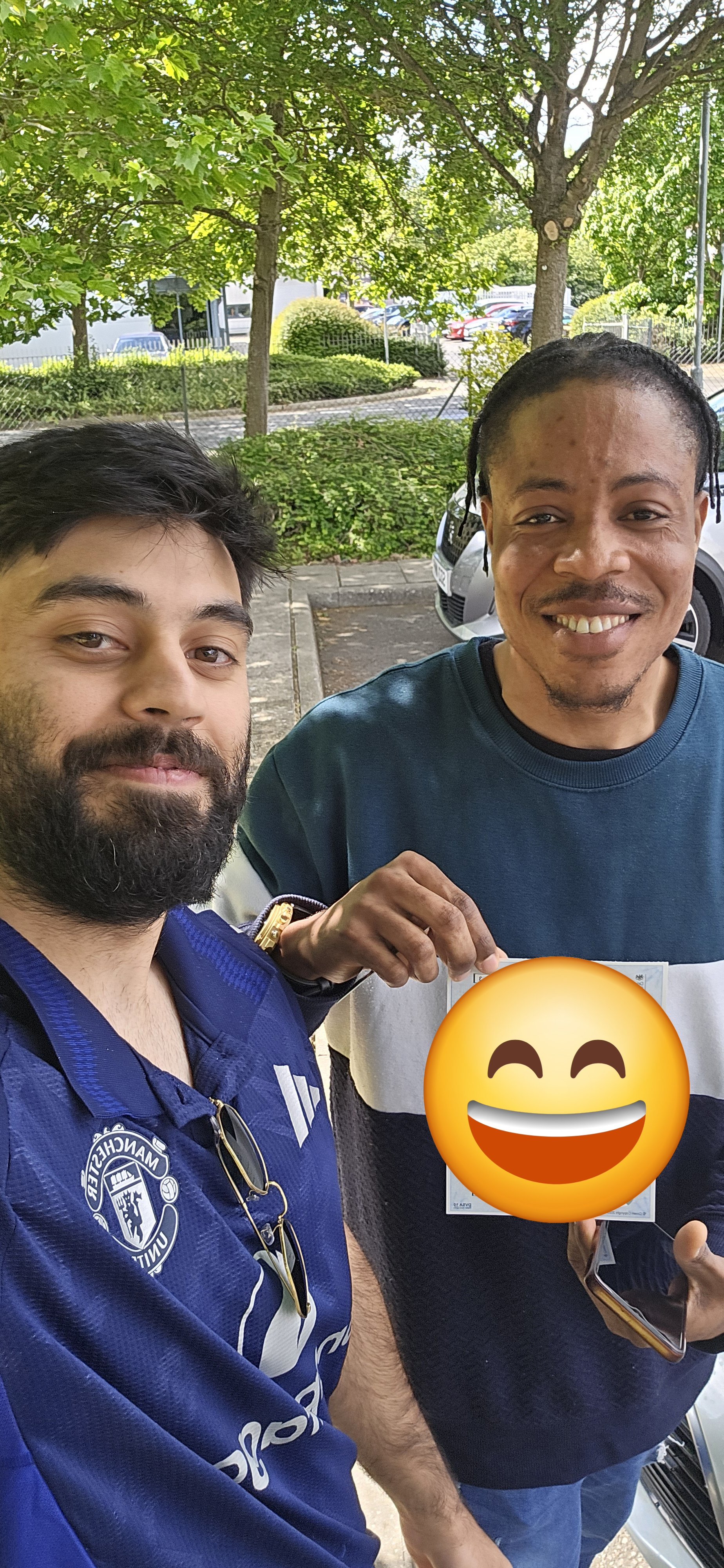 Two men taking a selfie outdoors, smiling. One is wearing a Manchester United jersey, the other is holding a smartphone and a paper with a big smiling emoji overlay. They are standing on a sidewalk with trees and parked cars in the background.
