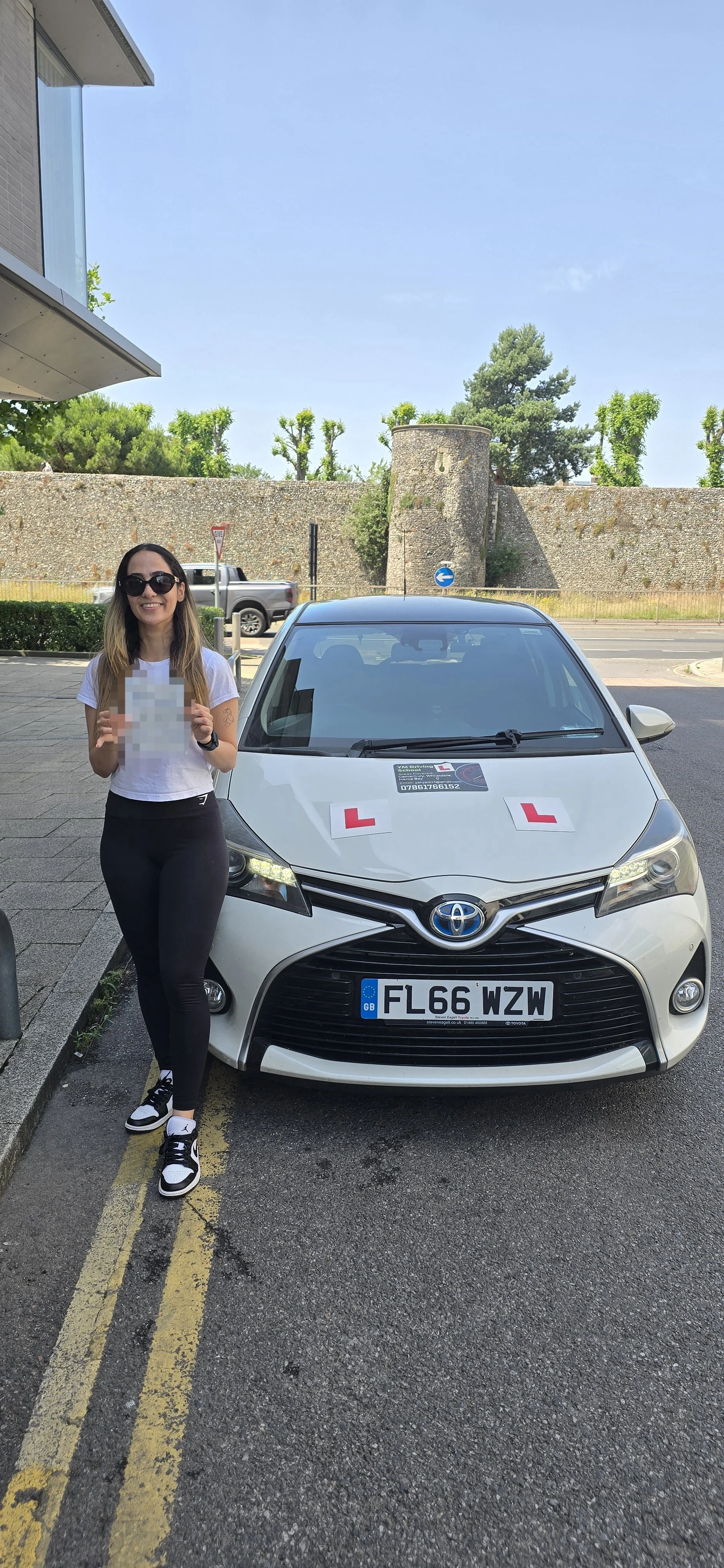 A smiling woman wearing sunglasses, a white t-shirt, and black pants standing next to a silver Toyota car with learner driver signs. She is holding a blurred document or object. The car has a UK license plate and is parked on the street with historic