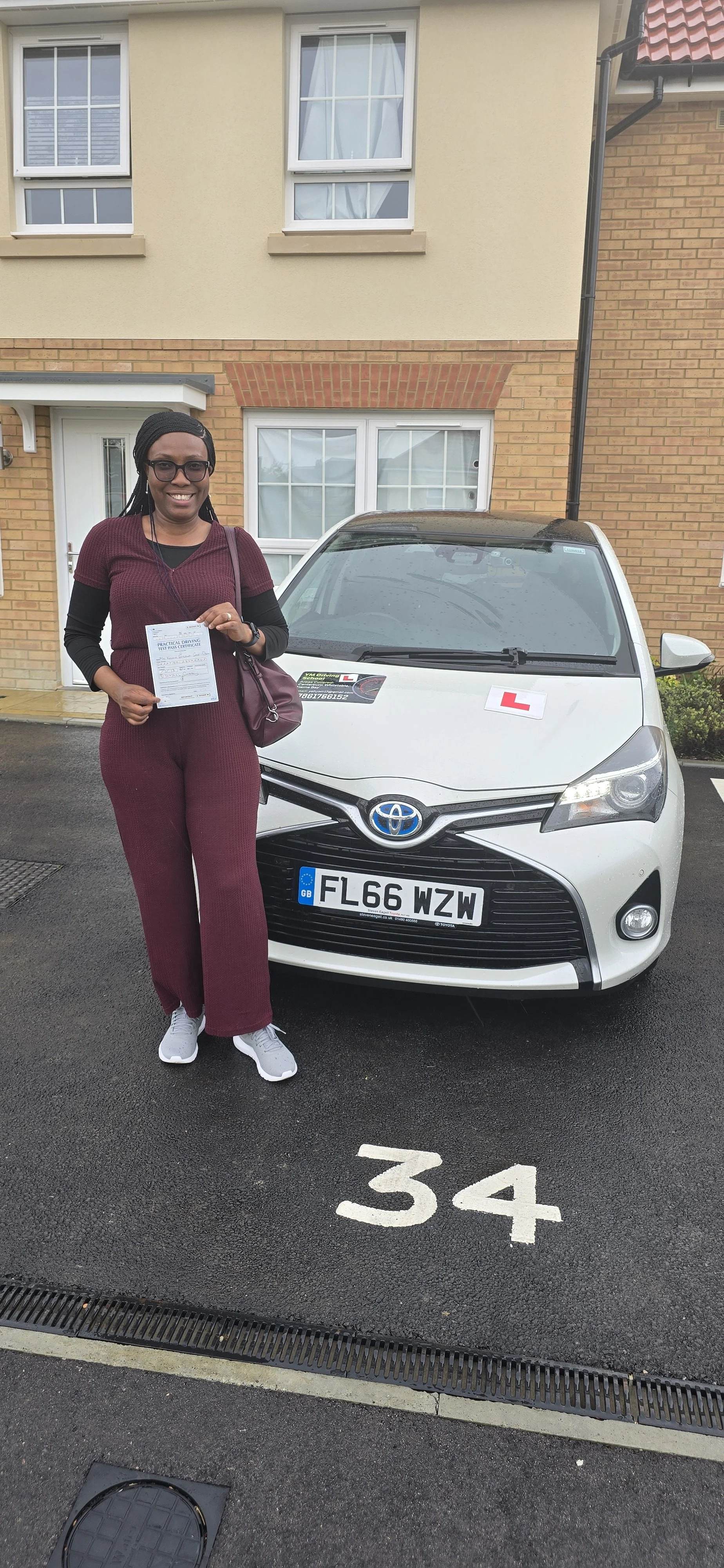 A smiling woman holding a driving test certificate standing next to a white Toyota car with 'L' plates, parked in space 34 outside a residential building.