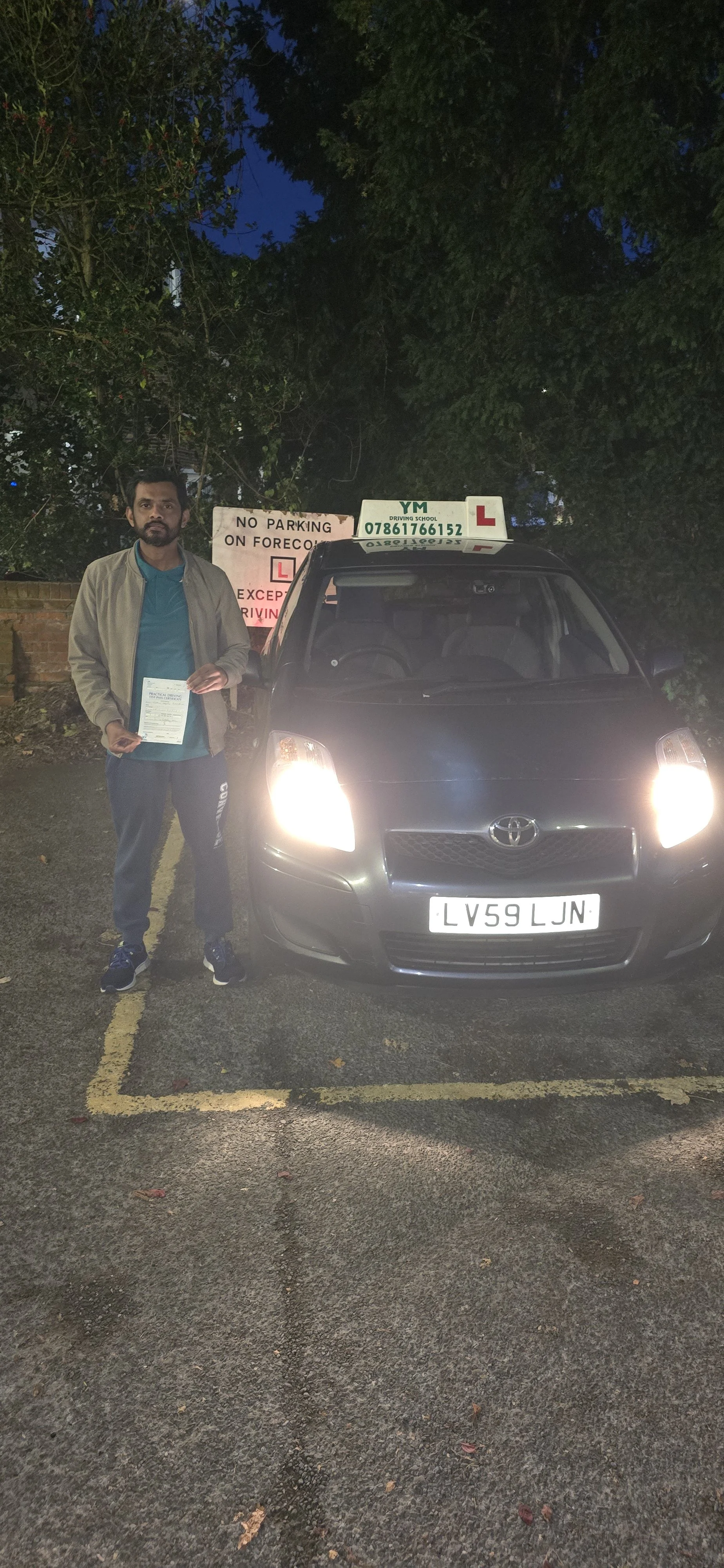 A man standing next to a black Toyota car holding a driving license, in a parking lot with a sign in the background that reads 'NO PARKING ON FORESOC' and a driving school sign on the car indicating it's a learner vehicle.