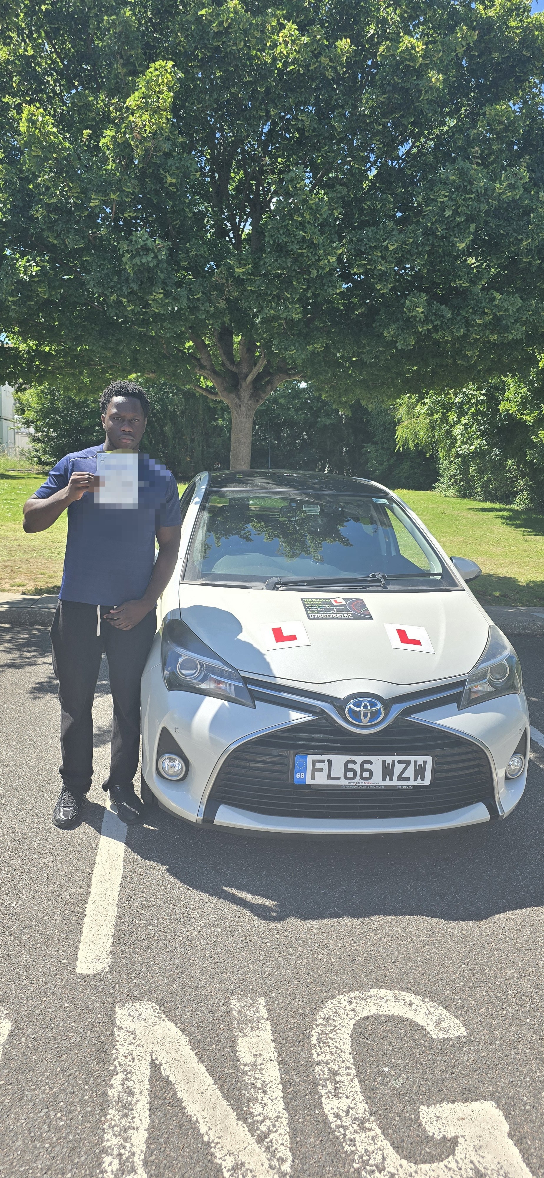Young man standing next to a white Toyota driving school car with learner 'L' plates parked in a marked parking space, holding a document, outdoors with a large green tree in the background.