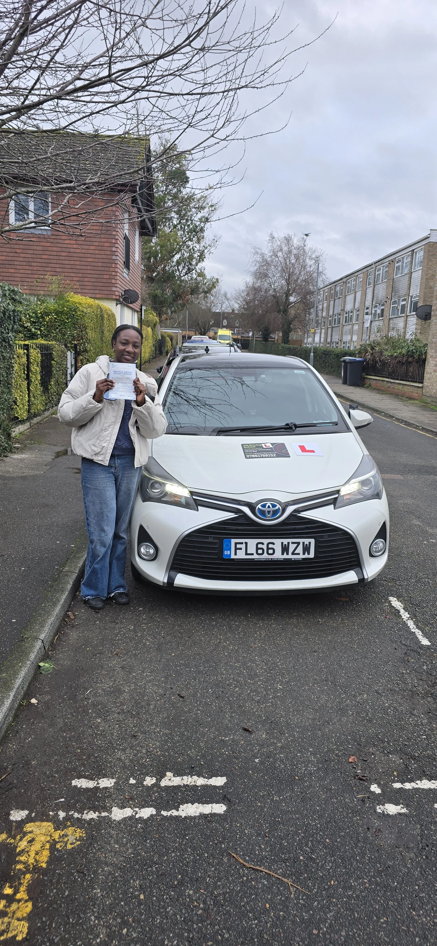 Woman holding a driver's license next to a white Toyota car with learner plates, parked on a residential street on a cloudy day.