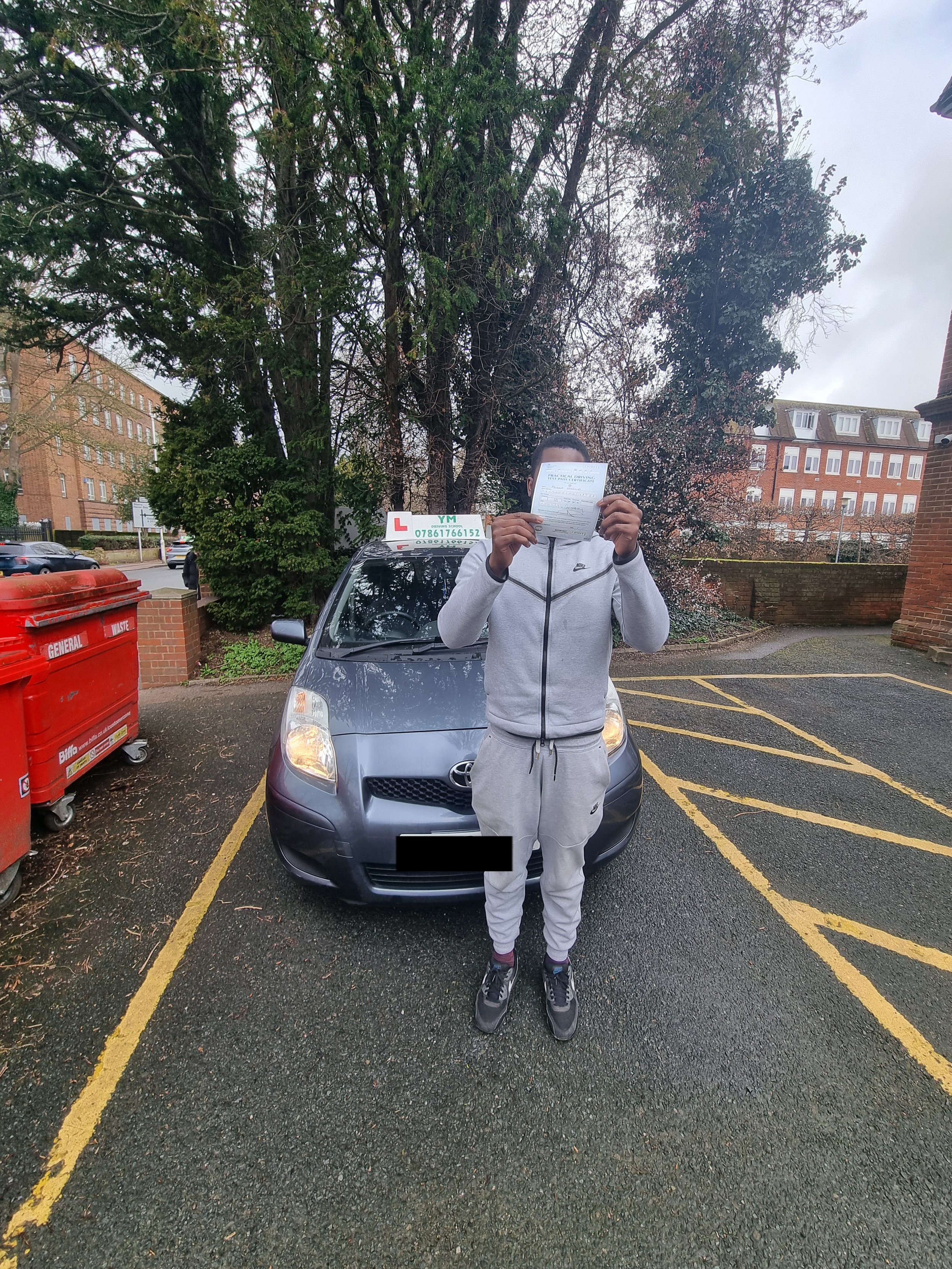 A man dressed in a grey tracksuit standing in front of a grey car, holding a piece of paper in front of his face. The car is parked in a marked parking space near trees and brick buildings.