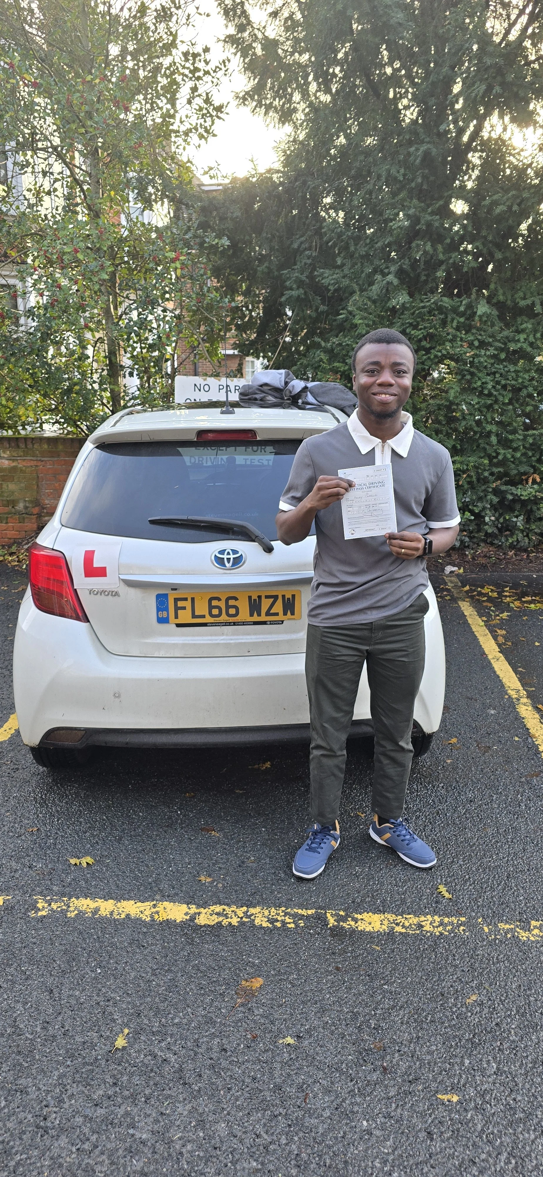 Young man standing in parking lot holding a driving test certificate, with a white Toyota car behind him. The car has a learner 'L' sign on the back, and the man is smiling. The parking lot has yellow lines and some fallen leaves, and there are trees