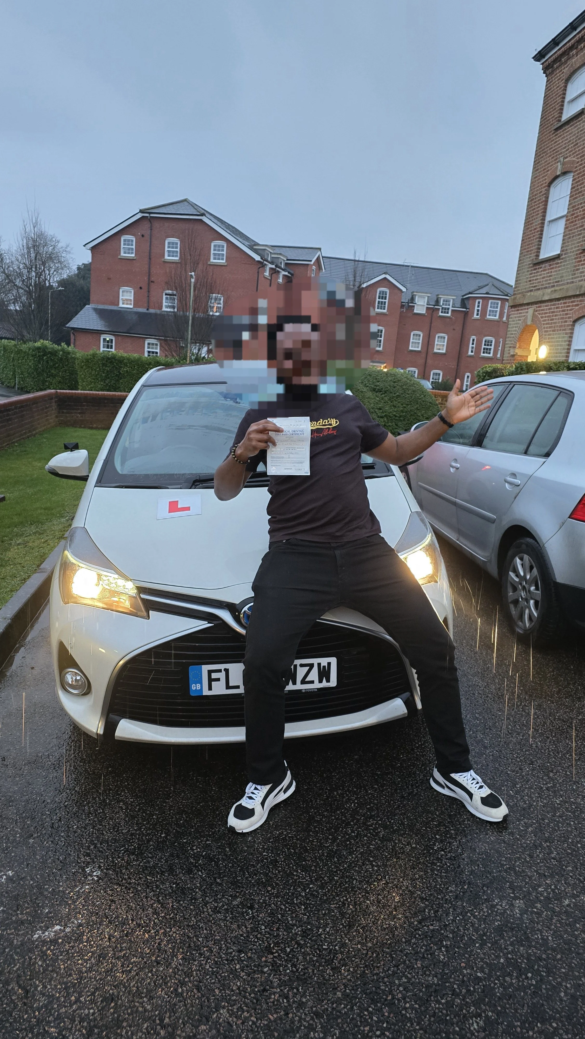 Person standing in front of a white car holding a document, with a blurred face, in a parking lot during dusk with other cars and red brick buildings in the background.