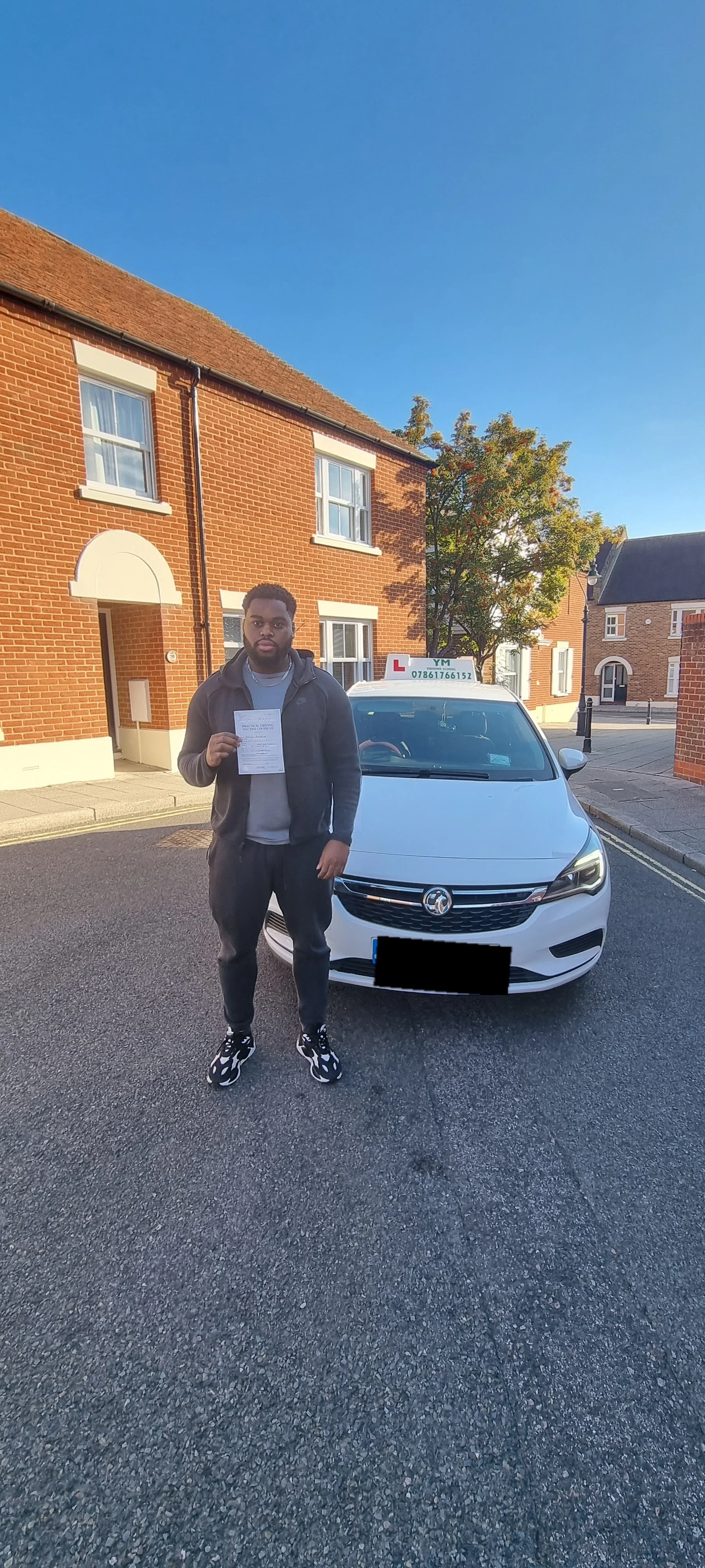 A man standing in front of a white driving school car with a learner's sign on top, holding a document, in a suburban neighborhood with red brick houses and a clear blue sky.