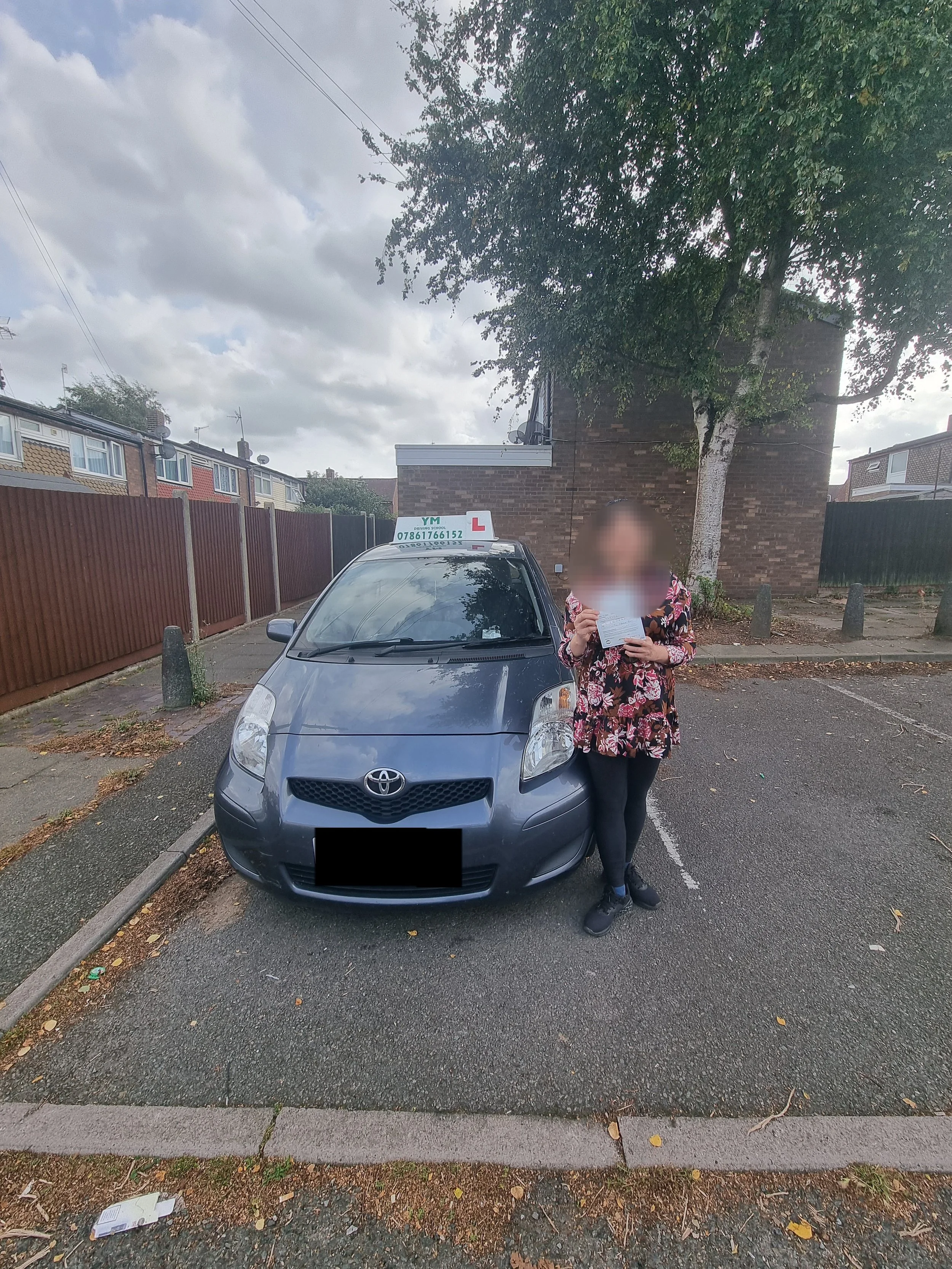 Person holding a paper standing next to a gray Toyota car with a learner driver sign on top in a parking lot.
