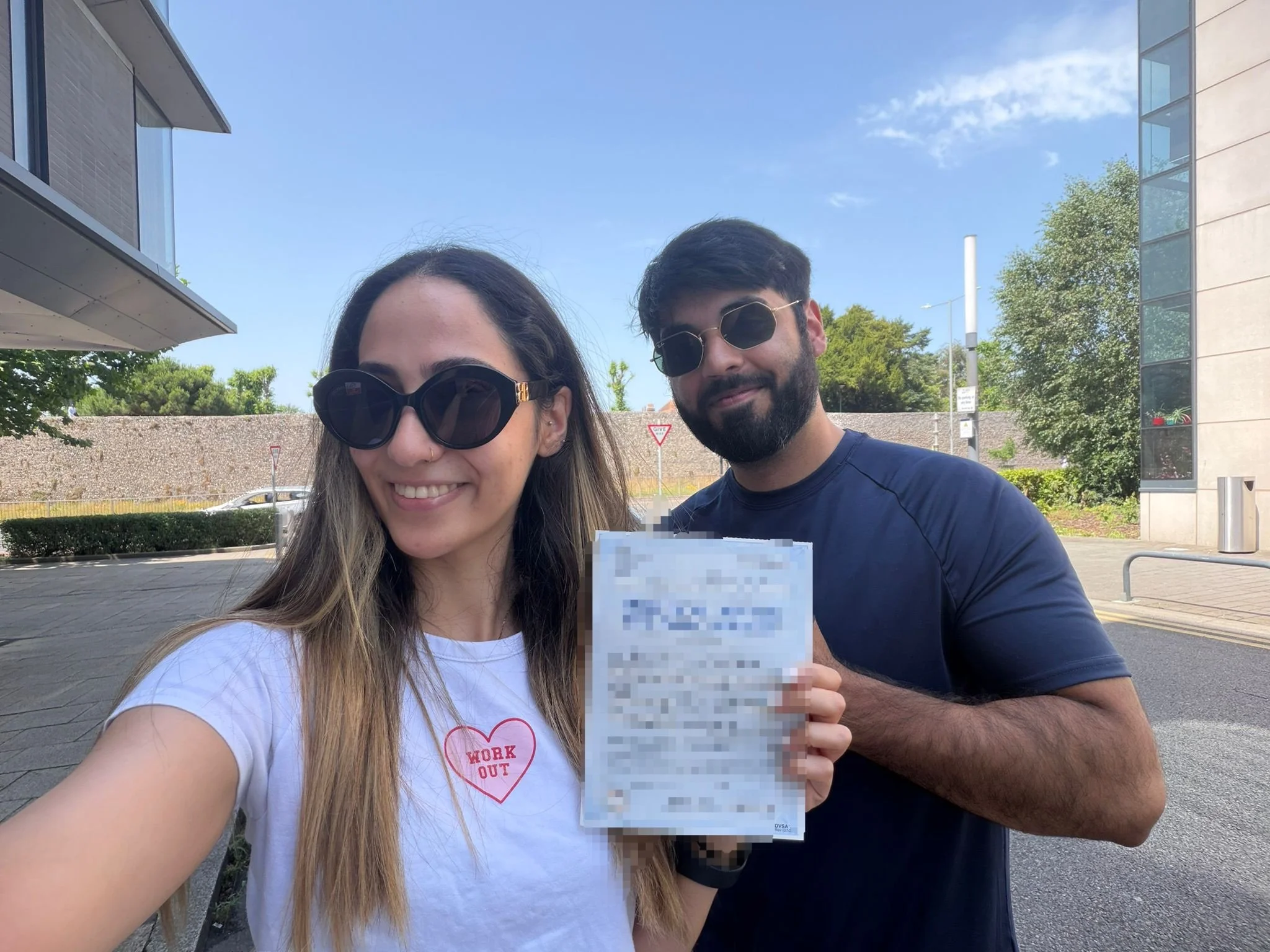 A woman and a man taking a selfie outdoors on a sunny day. The woman has long hair, wearing large sunglasses and a white t-shirt with a heart and words. The man has dark hair, a beard, and is wearing sunglasses and a navy shirt. They are smiling and 