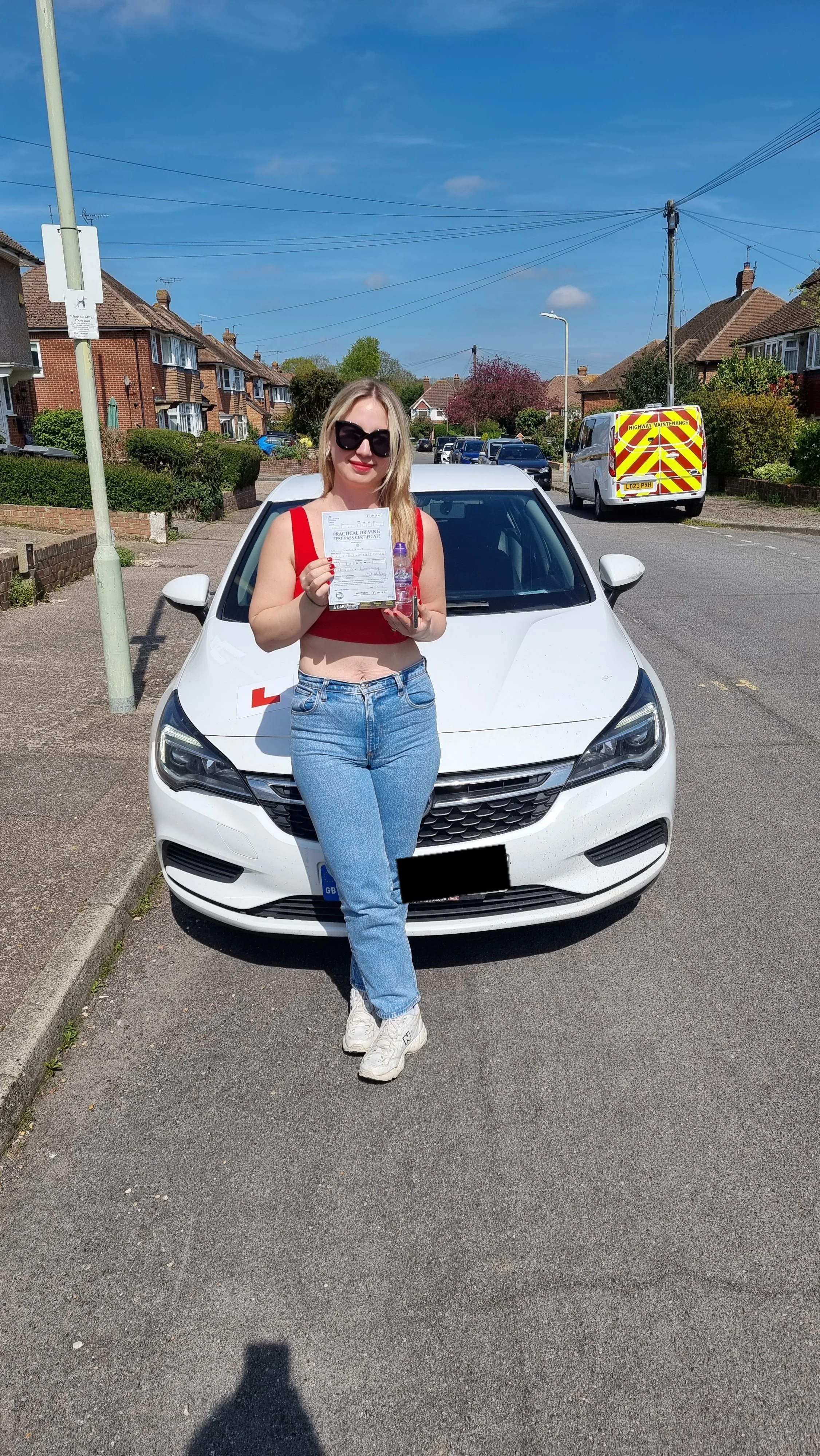 A young woman standing in front of a white car on a residential street, holding a practical driving test certificate and a water bottle, wearing sunglasses, a red crop top, blue jeans, and white sneakers, on a sunny day.