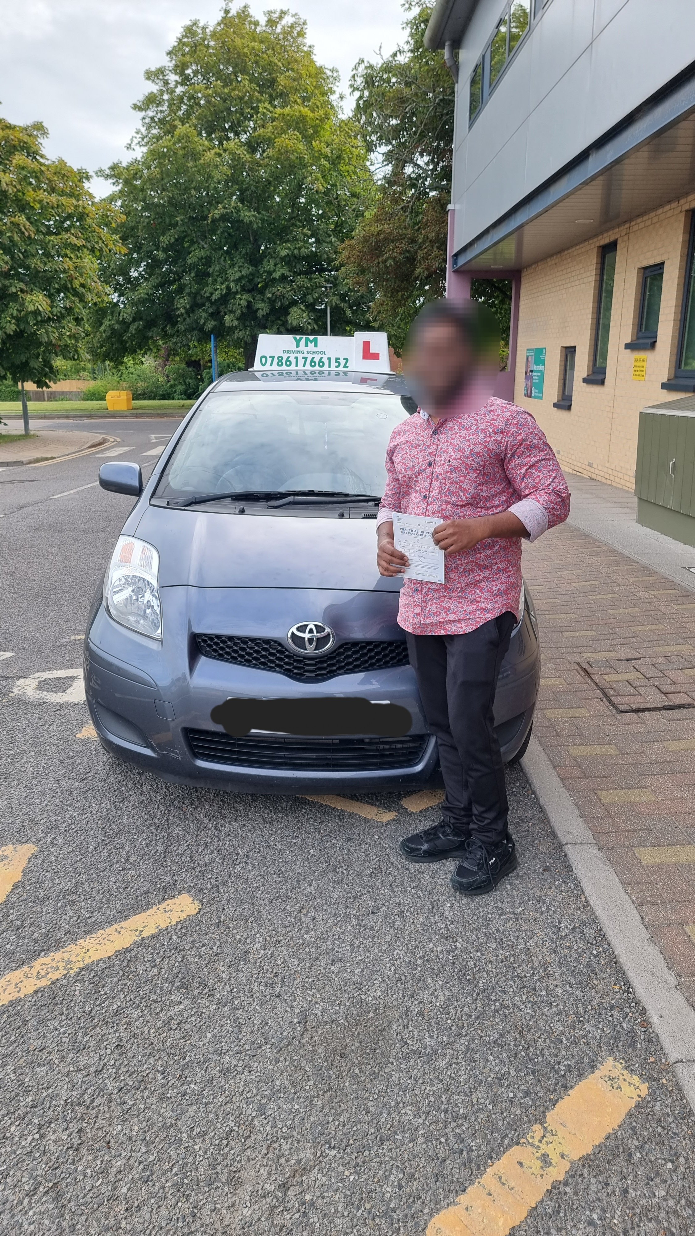 A man standing next to a grey Toyota driving school car holding a paper, with a sign on the roof indicating a driving instructor and learner license.