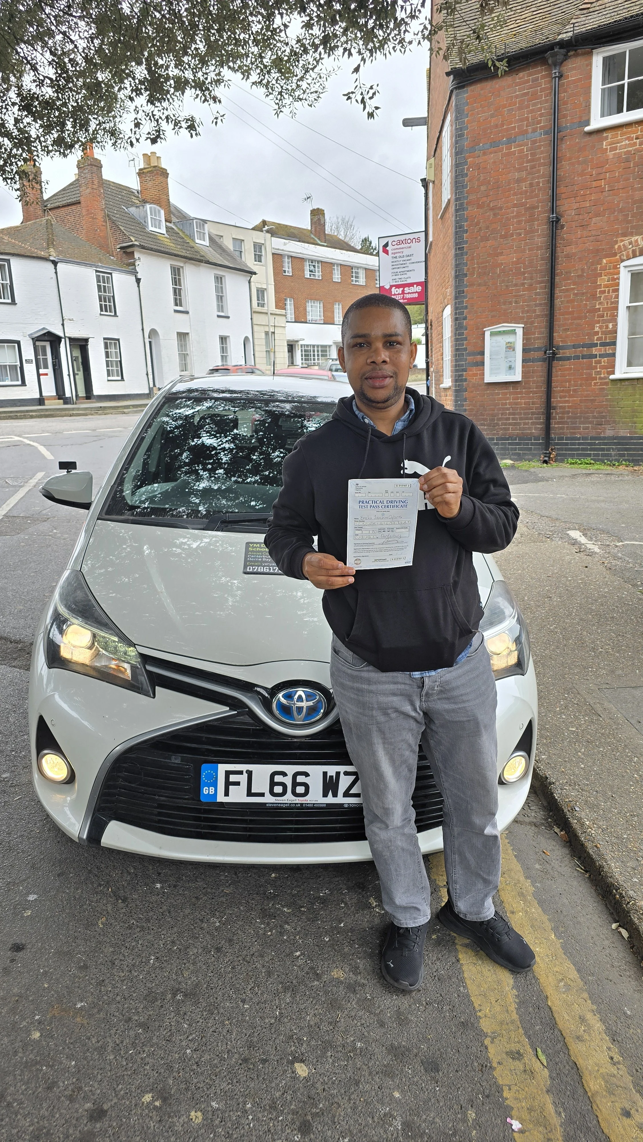 A man standing in front of a white car, holding a driving test pass certificate, on a city street with houses and a "for sale" sign in the background.