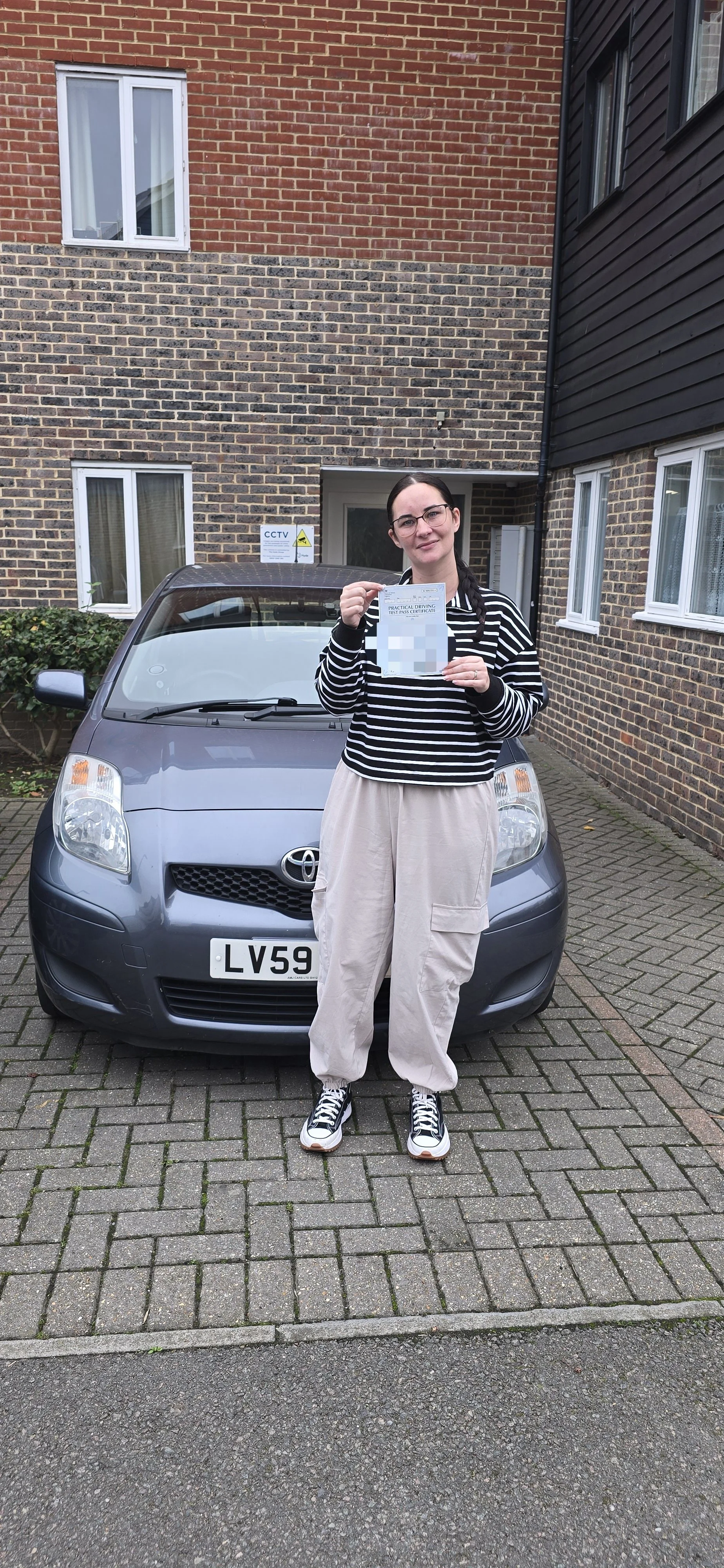 A woman with glasses and dark hair in pigtails standing in front of a gray car, holding a piece of paper, in a residential parking area with brick buildings around.