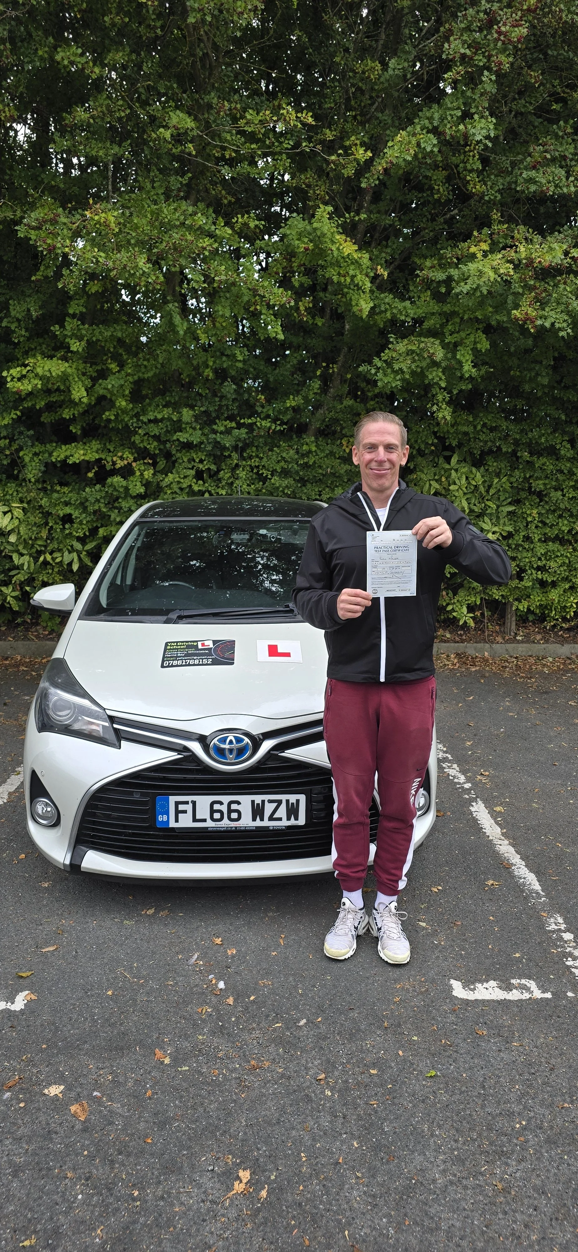 Man standing in parking lot holding driving test pass certificate, dressed in athletic wear, in front of a white Toyota car with learner 'L' signs, green foliage background.