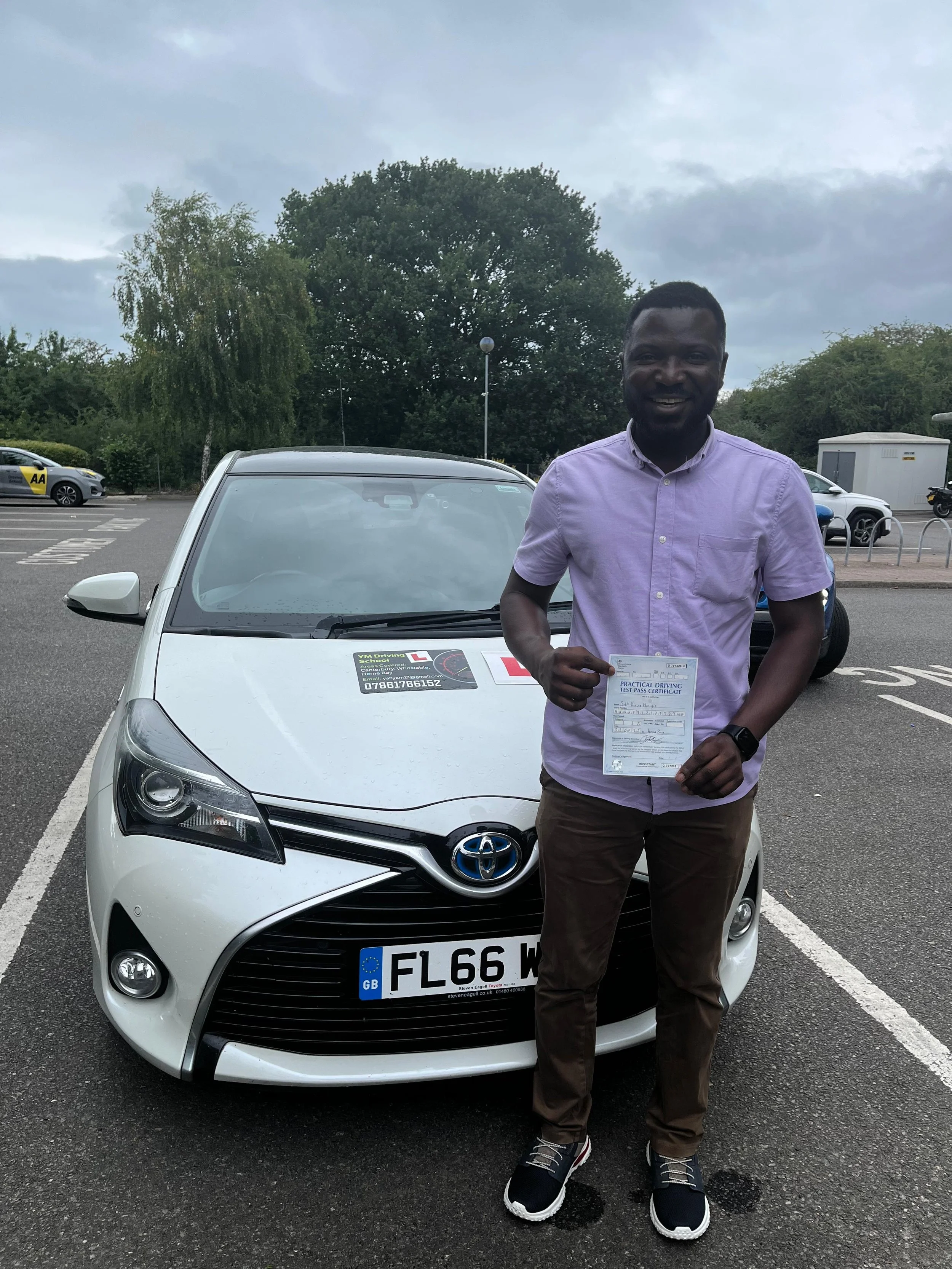 A man standing in front of a silver Toyota car holding a driving certificate, smiling, in a parking lot with trees and other cars in the background.