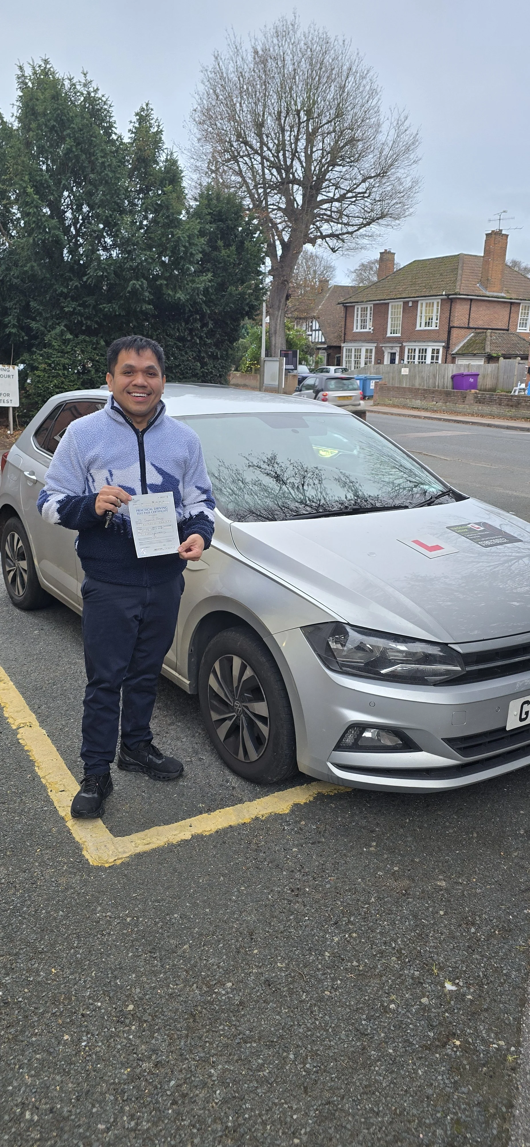 A smiling man holding a driving test certificate stands next to a silver car on a street with residential houses in the background.