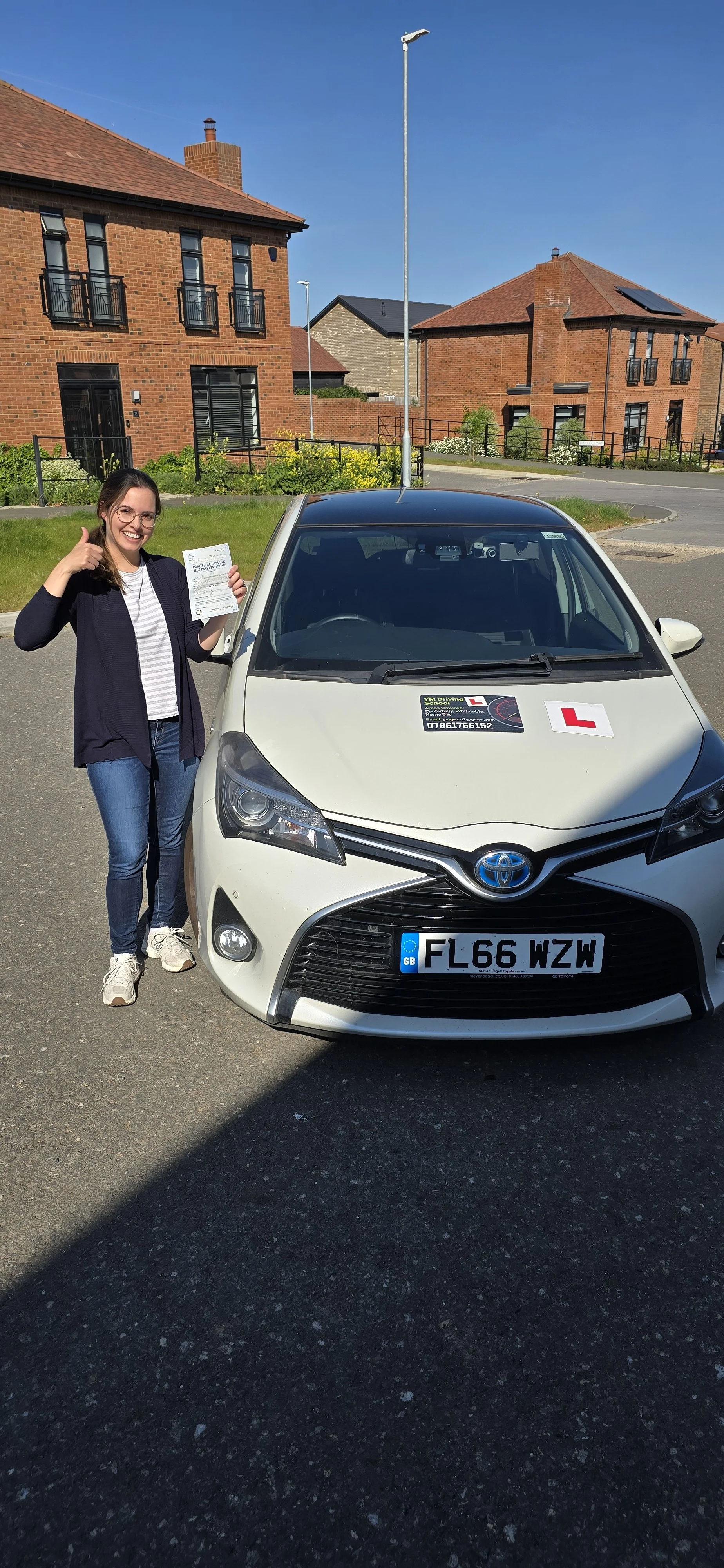 A young woman standing next to a white Toyota driving school car, holding a piece of paper and giving a thumbs-up. The car has an 'L' learner driver sign and a UK license plate, parked on a residential street with brick houses in the background under