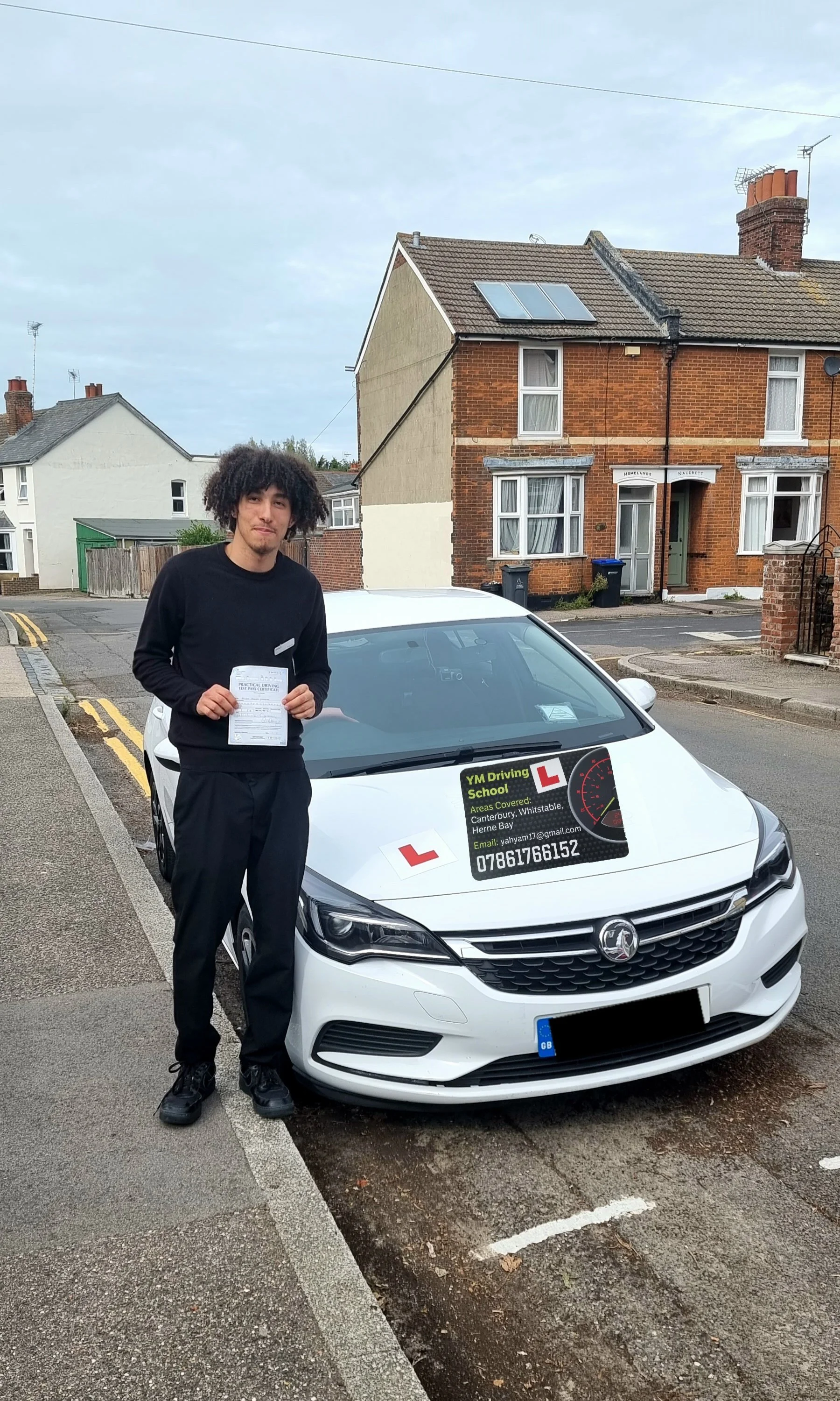 Young man holding a driving test sheet in front of a white car marked with learner driver signs, parked on a suburban street.