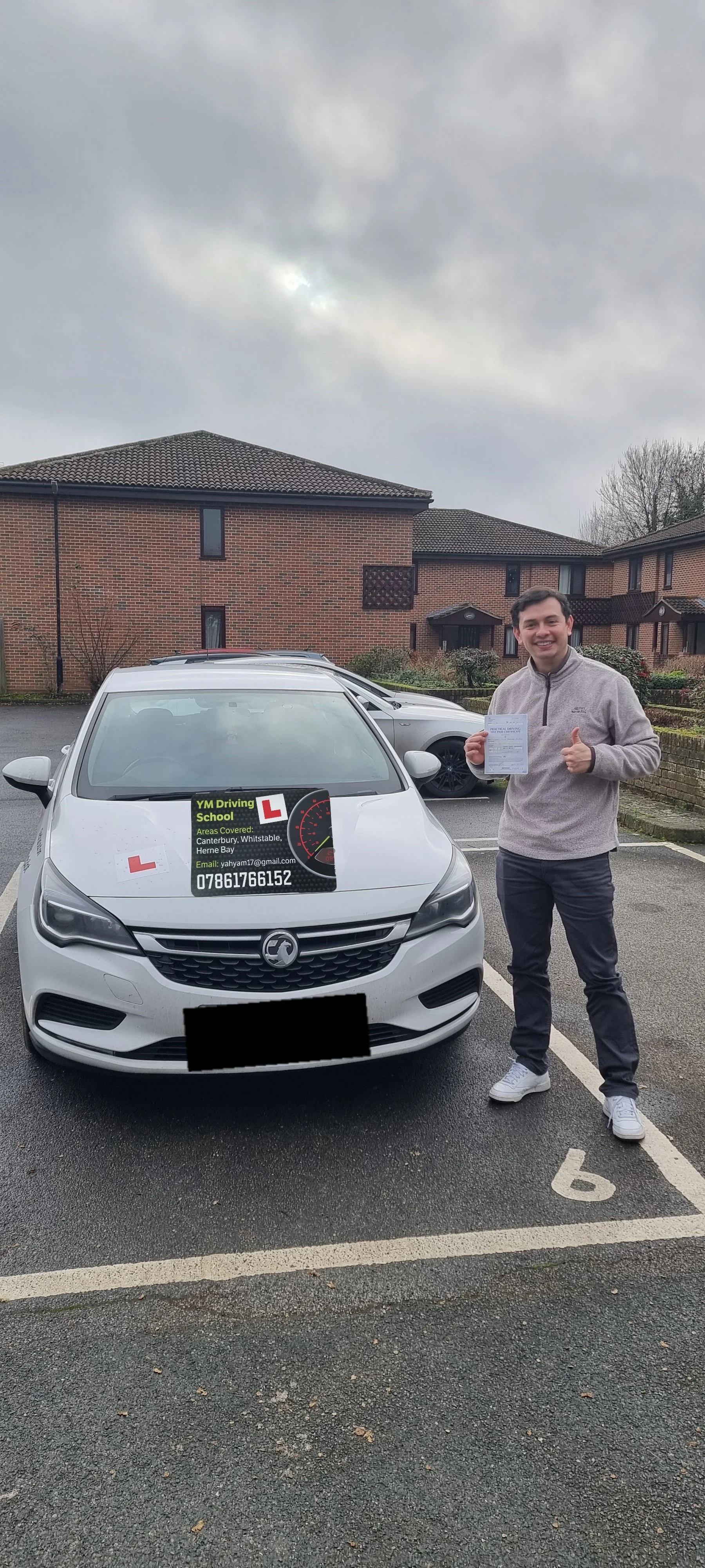 A young man standing next to a white car in a parking lot, holding a piece of paper and giving a thumbs up, with a sign on the car advertising a driving school.