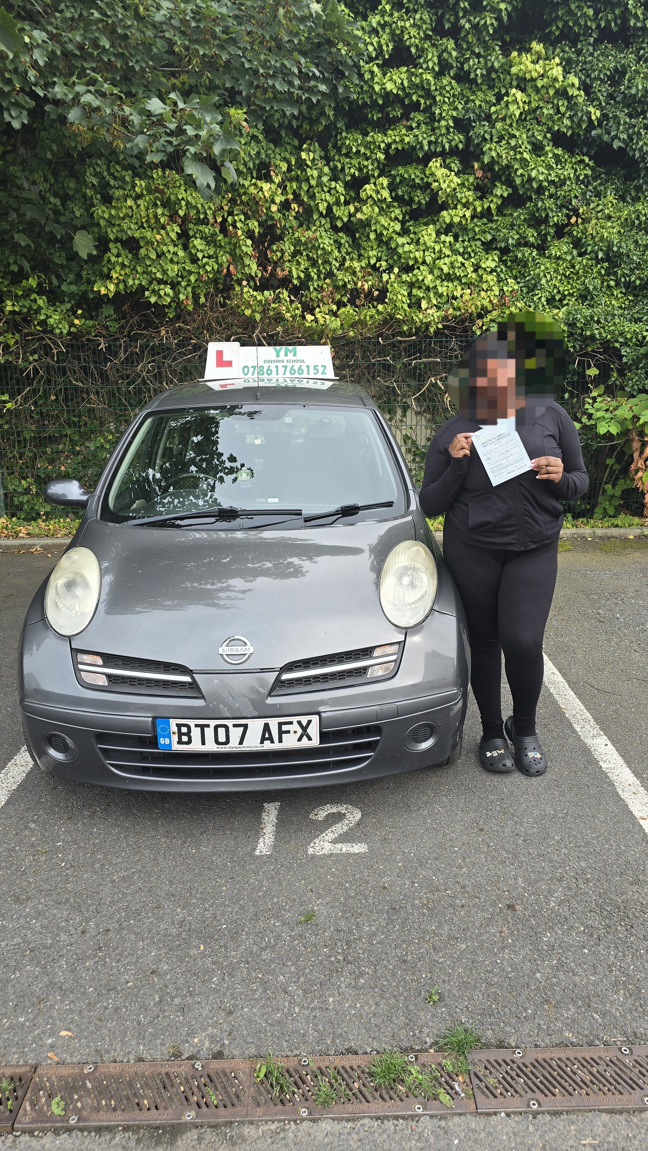 A woman standing next to a gray Nissan car with a driving school sign on top, holding a document, in a parking lot marked with the number 12, with green foliage in the background.