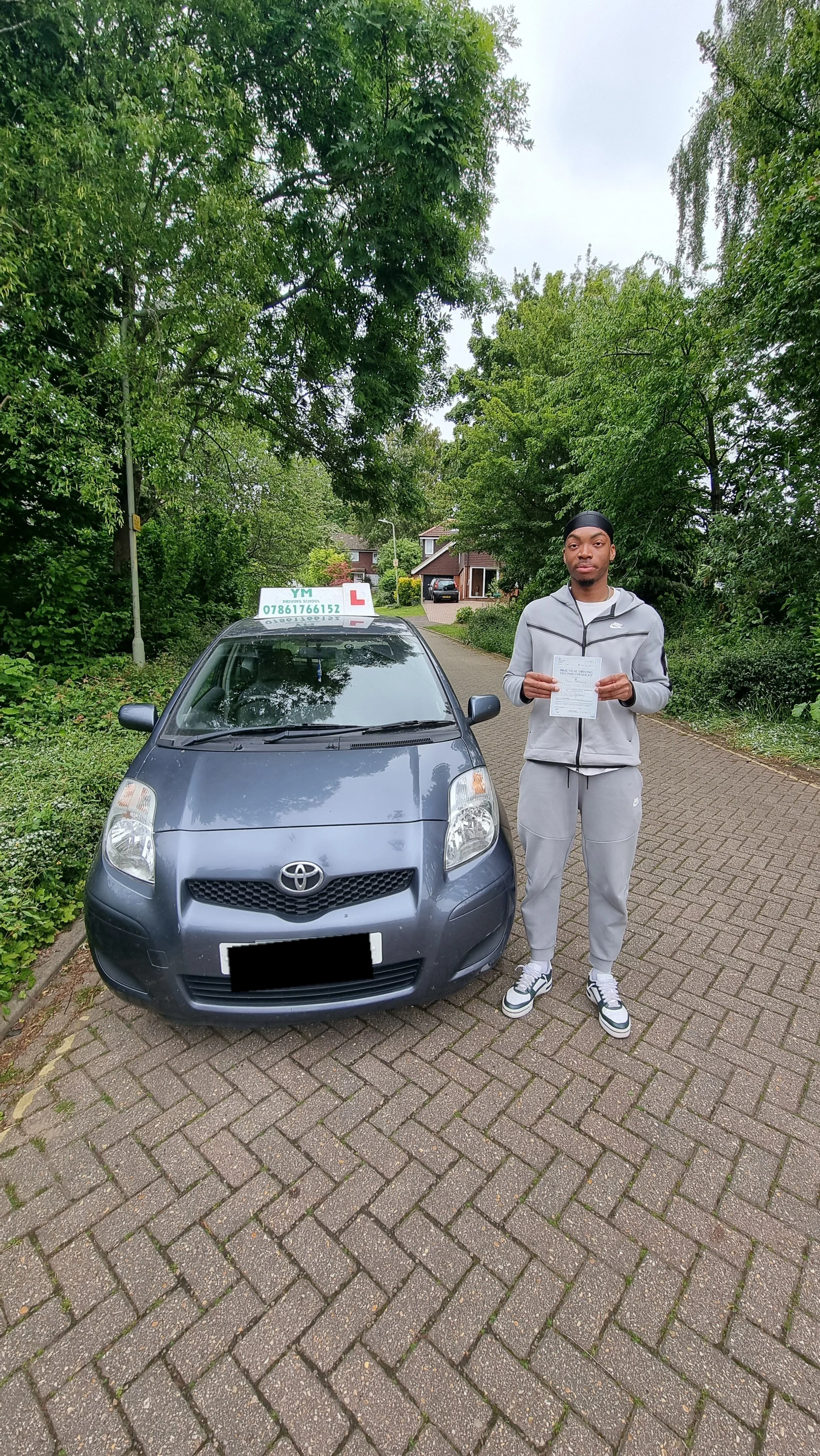 A young man in gray athletic wear holding a paper standing next to a dark gray Toyota car with a driving school sign on top, on a suburban street lined with trees and houses.