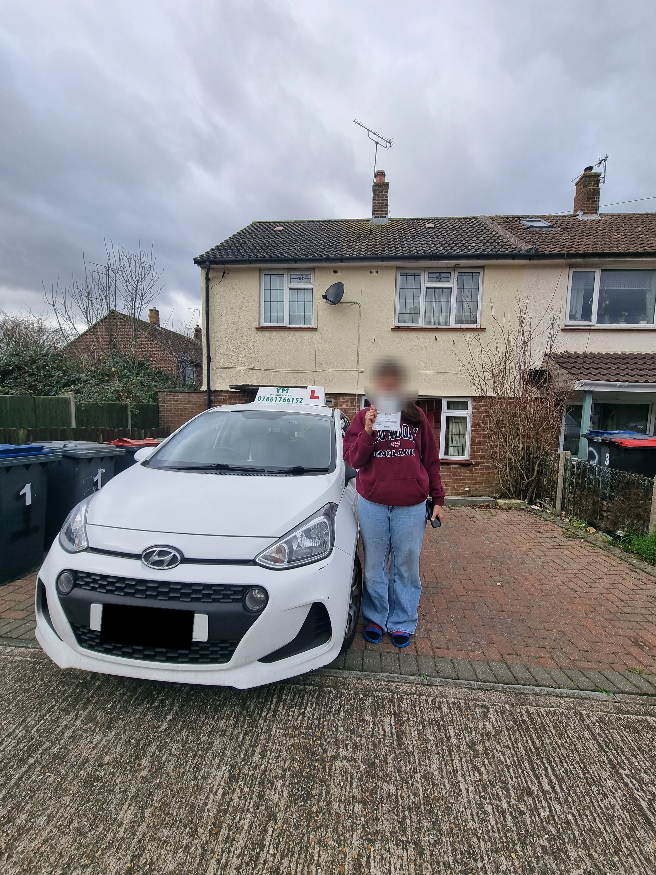 A person standing beside a white Hyundai driving school car in a residential driveway, holding a document.