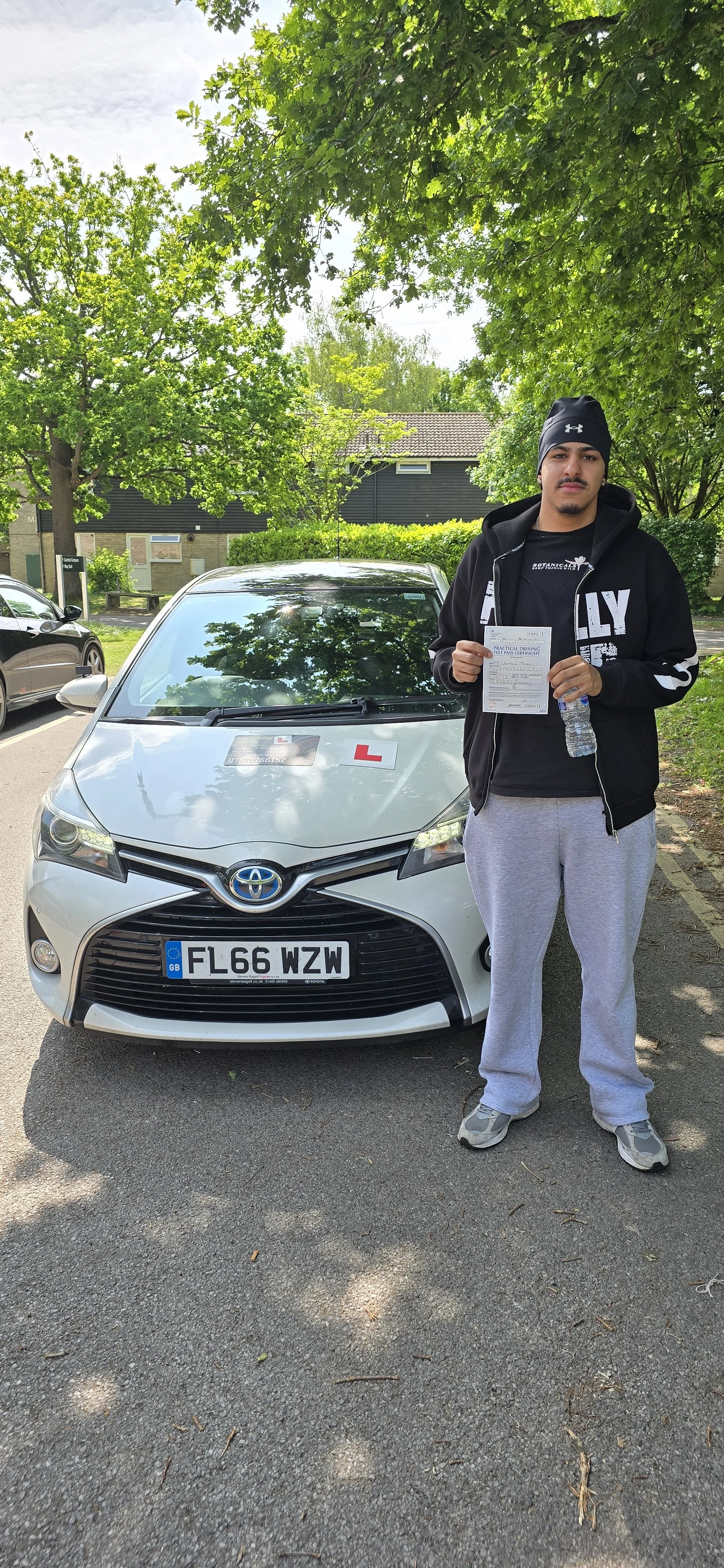 A young man standing outdoors next to a silver Toyota car holding a paper and water bottle, with driving instructor signs on the car's hood.