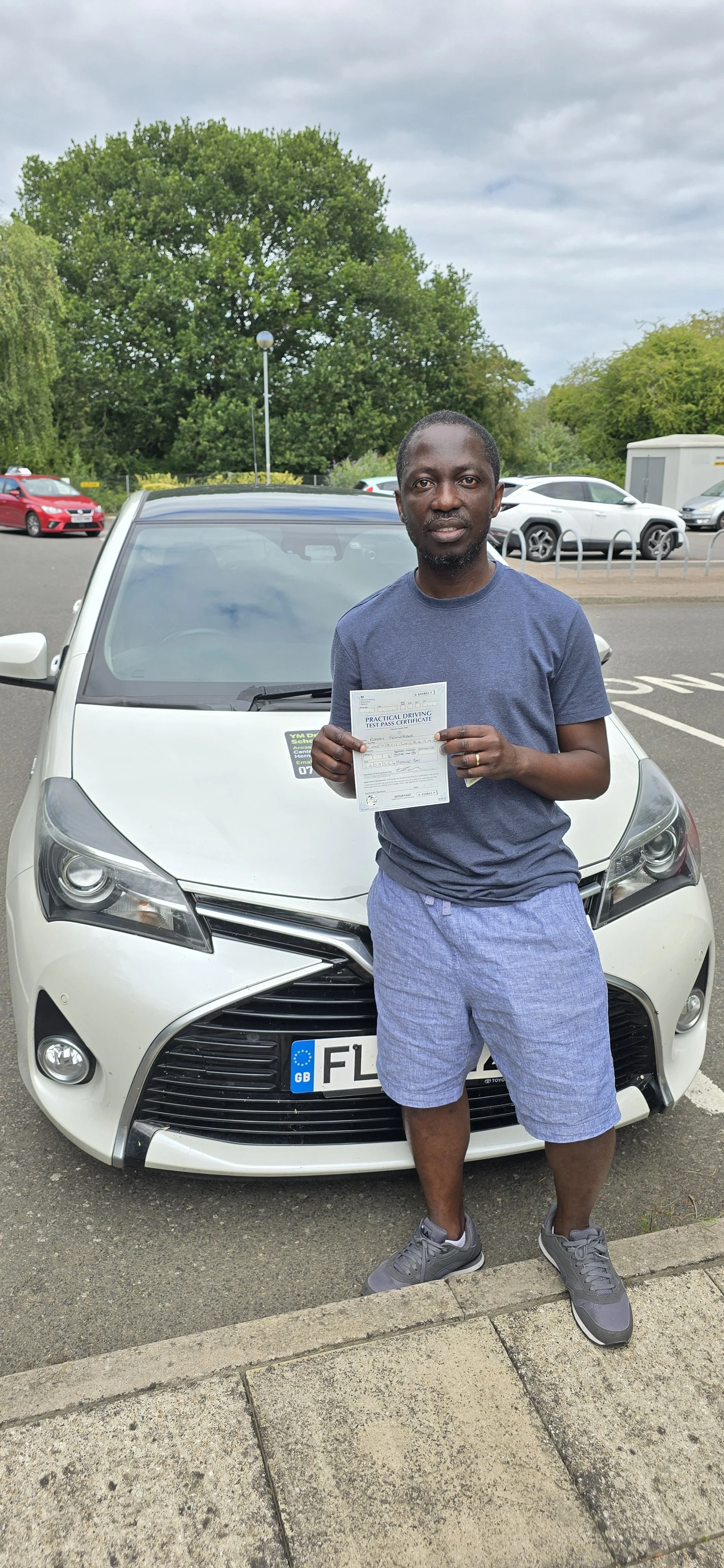 Man standing in front of a white car holding a provisional driving license, in an outdoor parking lot with green trees and cloudy sky.