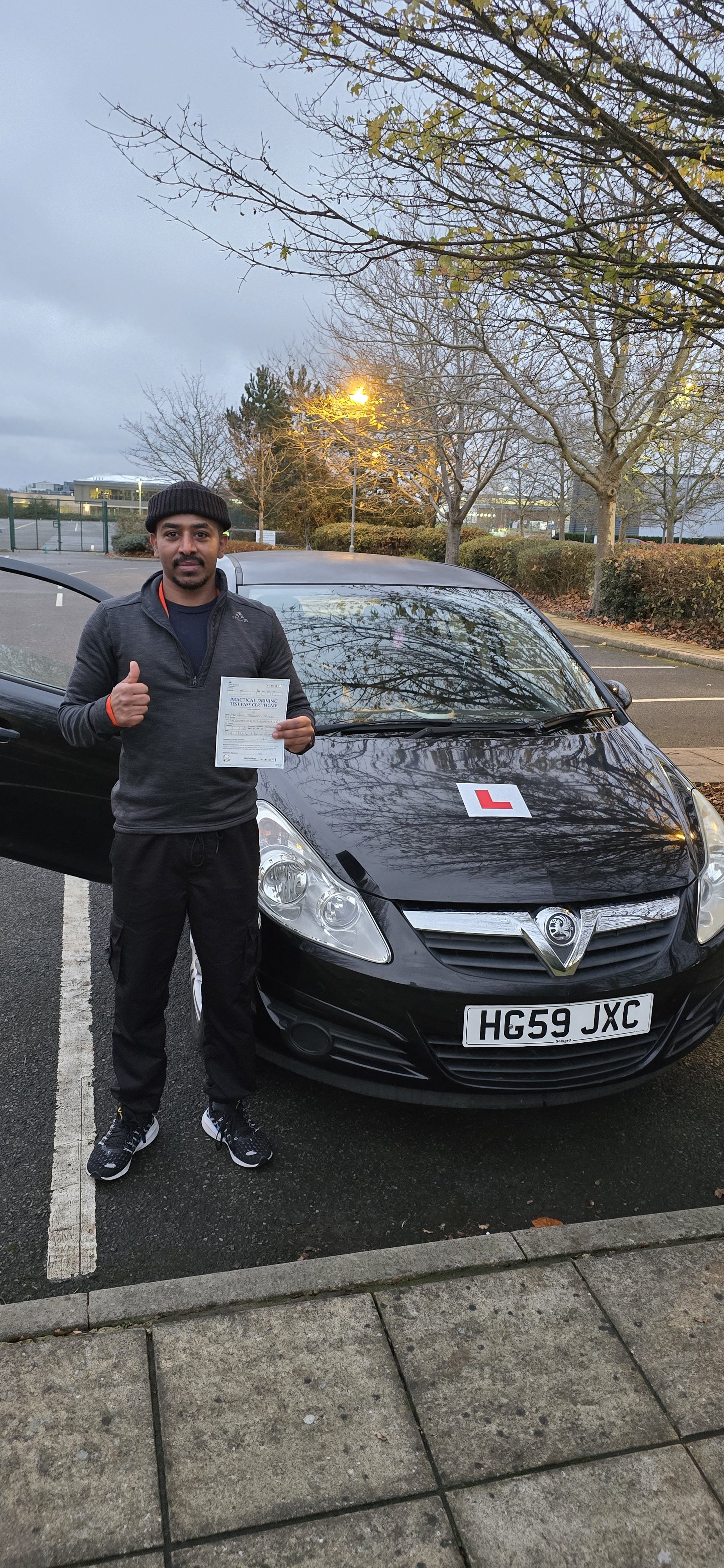 A man standing next to a black Vauxhall car in a parking lot, holding a certificate and giving a thumbs-up. The car has a learner driver 'L' sign on the hood. The man is dressed in black athletic clothing and a beanie, with trees and overcast sky in 