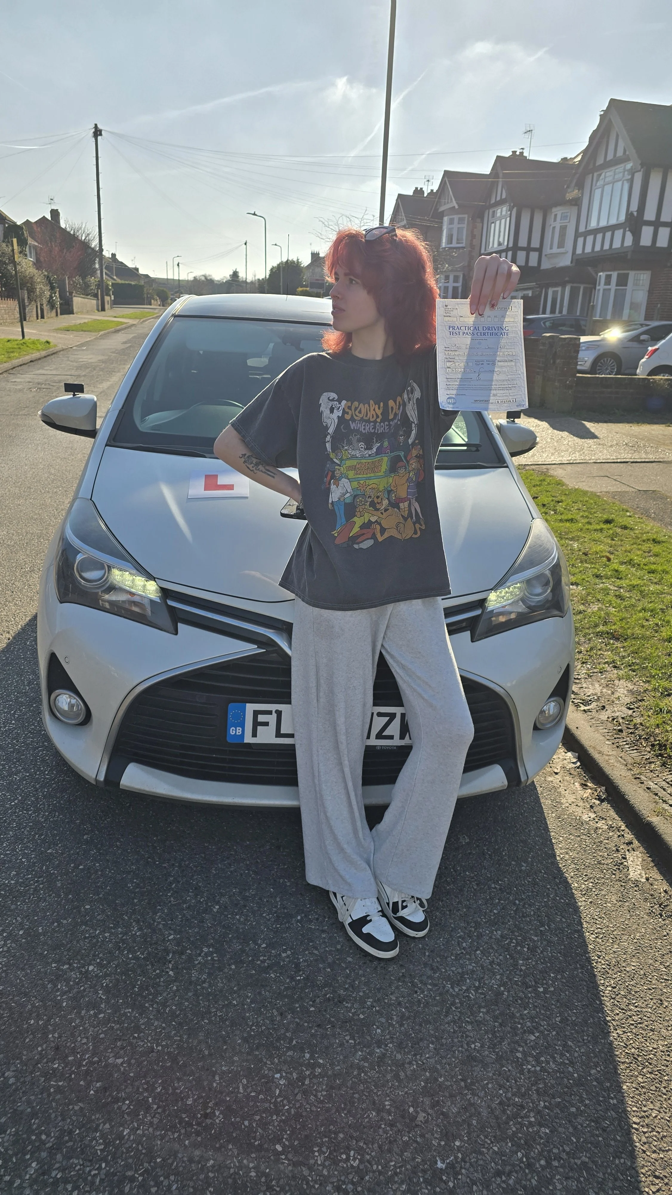 A woman with red hair standing in front of a white car with a learner driver sign on the hood, holding a driving test pass certificate in her right hand. She's wearing a Grateful Dead t-shirt and gray sweatpants, on a suburban street with houses and 