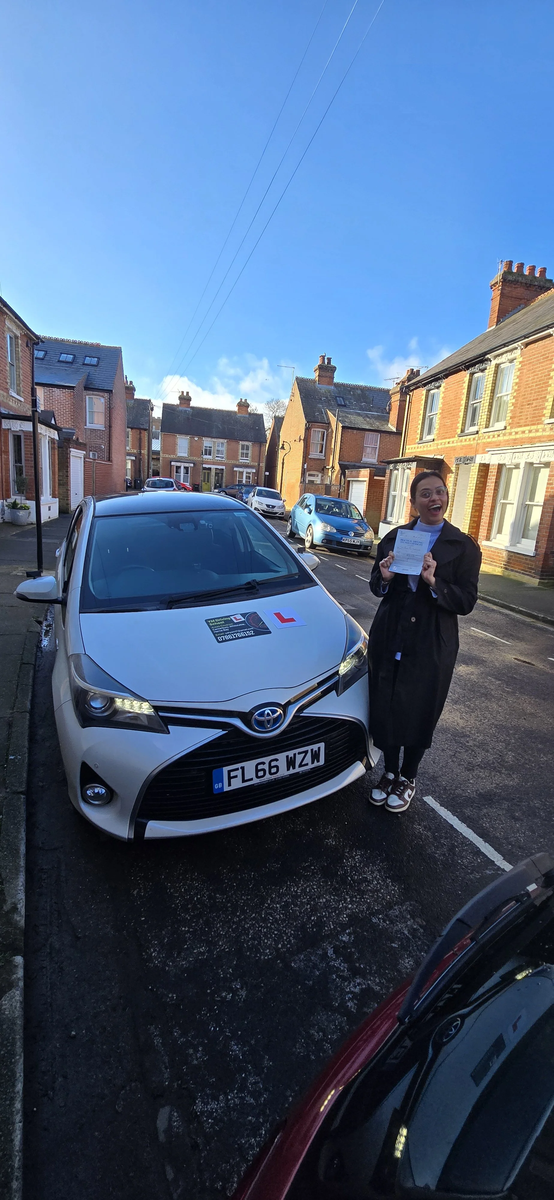 A woman holding a driving test completion certificate stands on a street next to a white Toyota car with an L-plate, indicating a learner driver. The woman is smiling, and there are parked cars and brick houses in the background under a clear blue sk