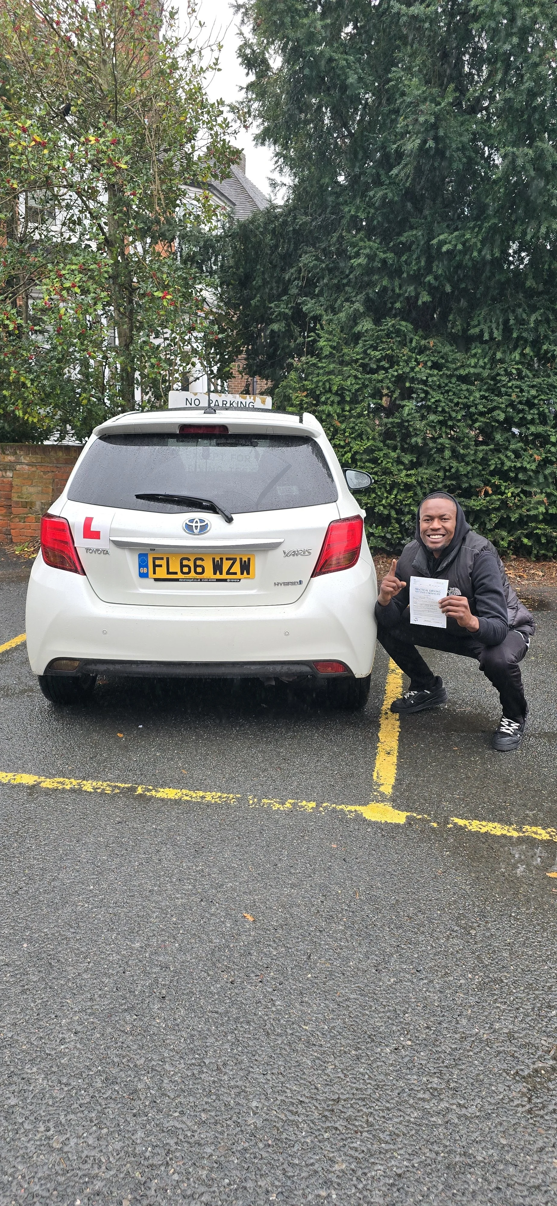 A man crouching next to a white Toyota car with a learner driver sign on top, in an outdoor parking lot on a wet day. The man appears happy and is holding a document, possibly a driving test result or license.