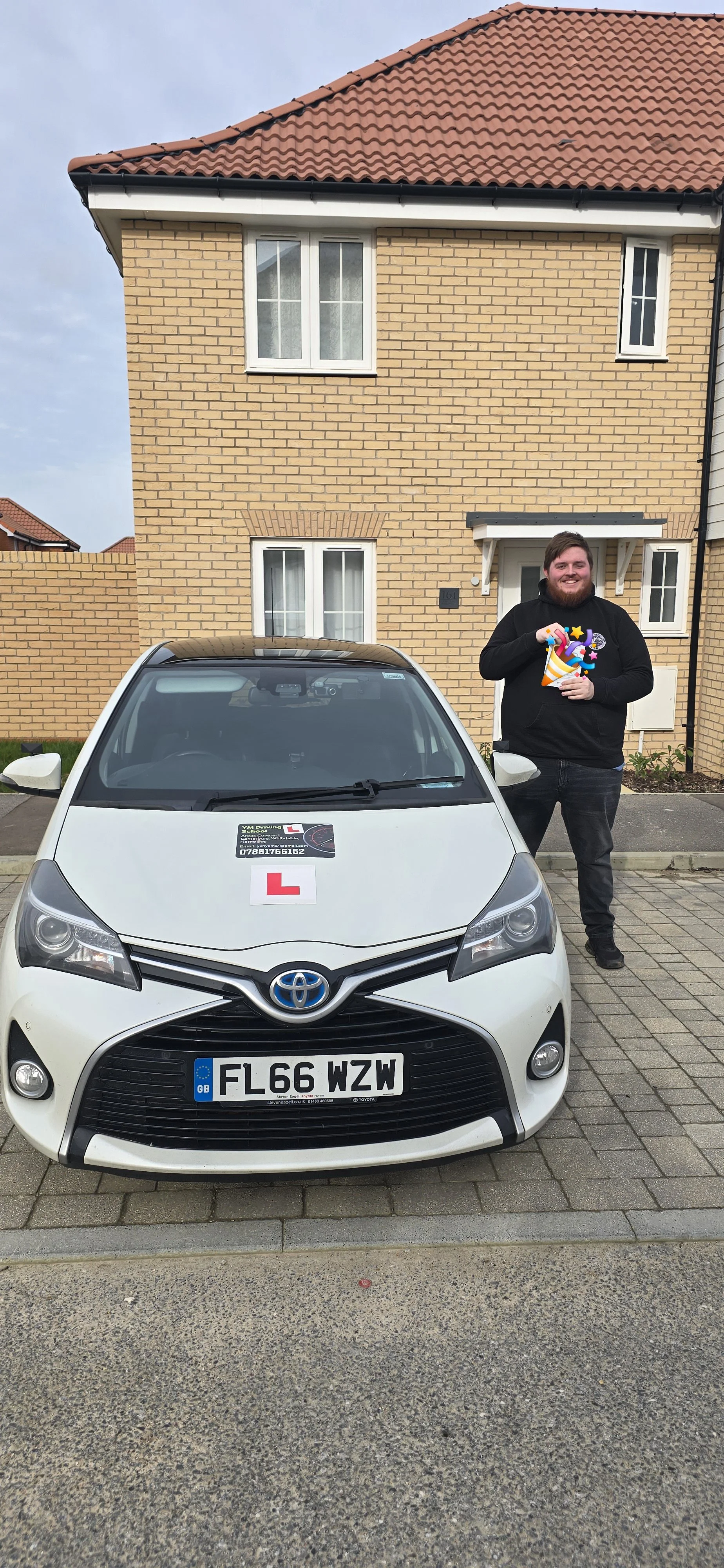 A man standing next to a white Toyota car with a learner driver sign on the hood, holding a colorful party horn, in front of a brick house with a tiled roof.
