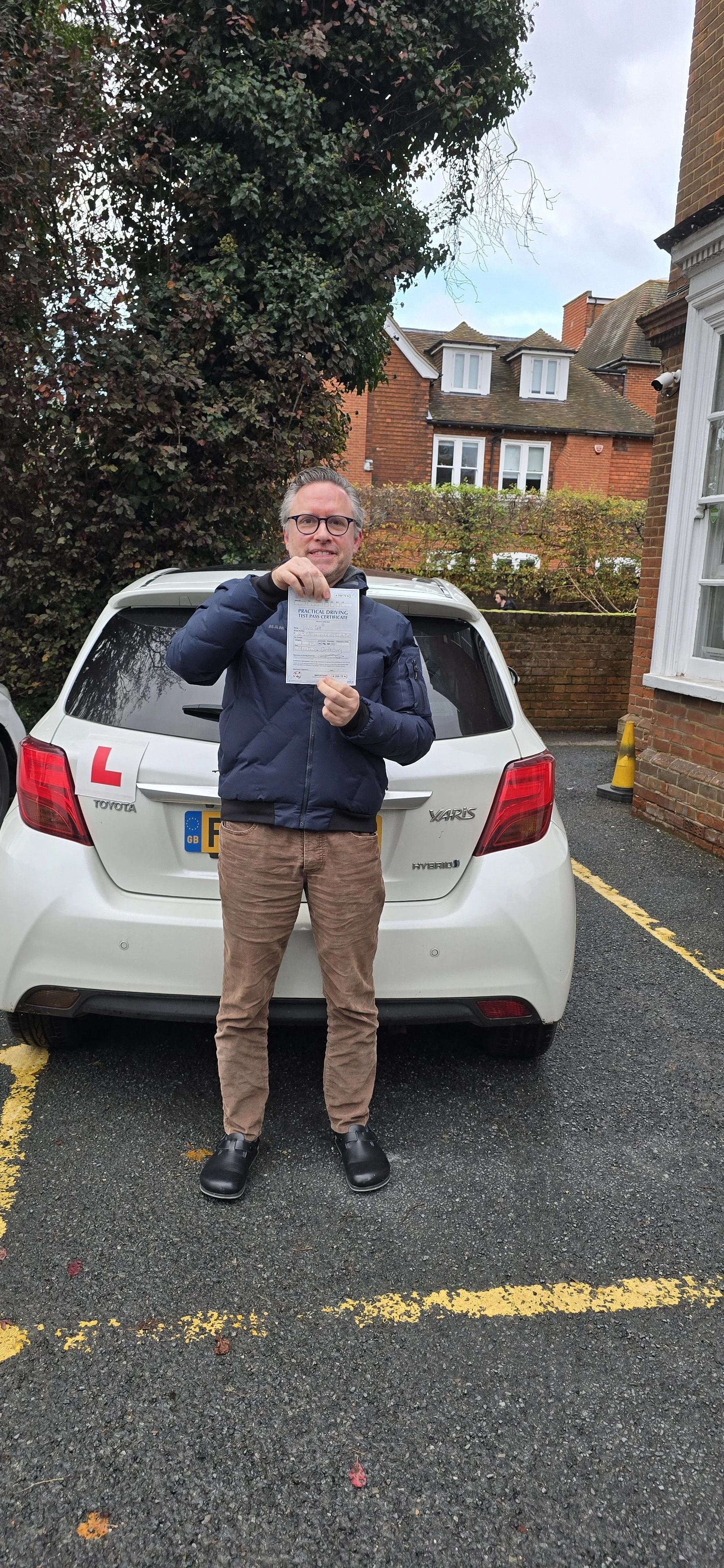 A man standing in front of a white Toyota Yaris hybrid car holding a practical driving test pass certificate, with an 'L' learner driver sign on the car, on a parking lot next to a brick building and trees.