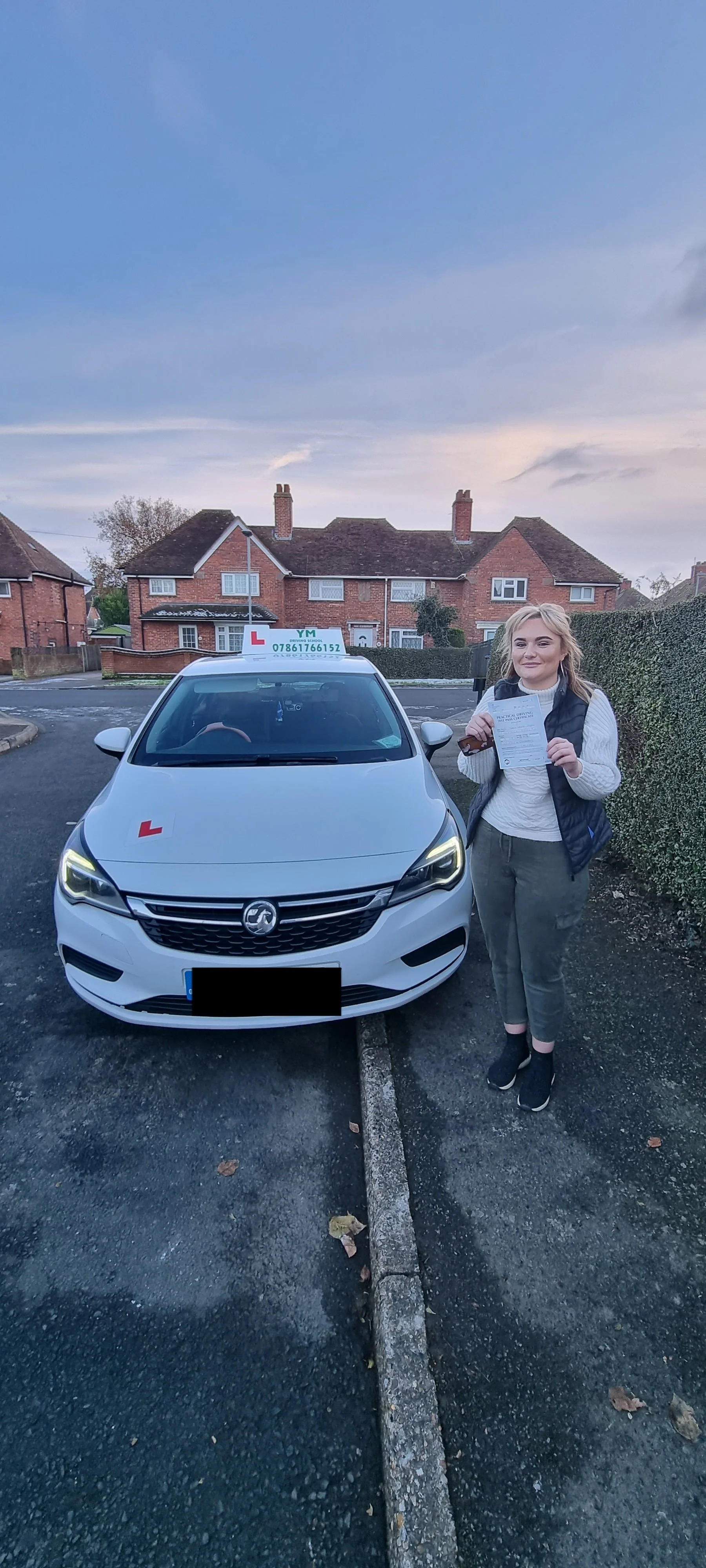 A woman standing next to a white driving school car with 'L' plates and a sign on top, holding a document and a small stuffed toy, in a residential neighborhood during dusk.