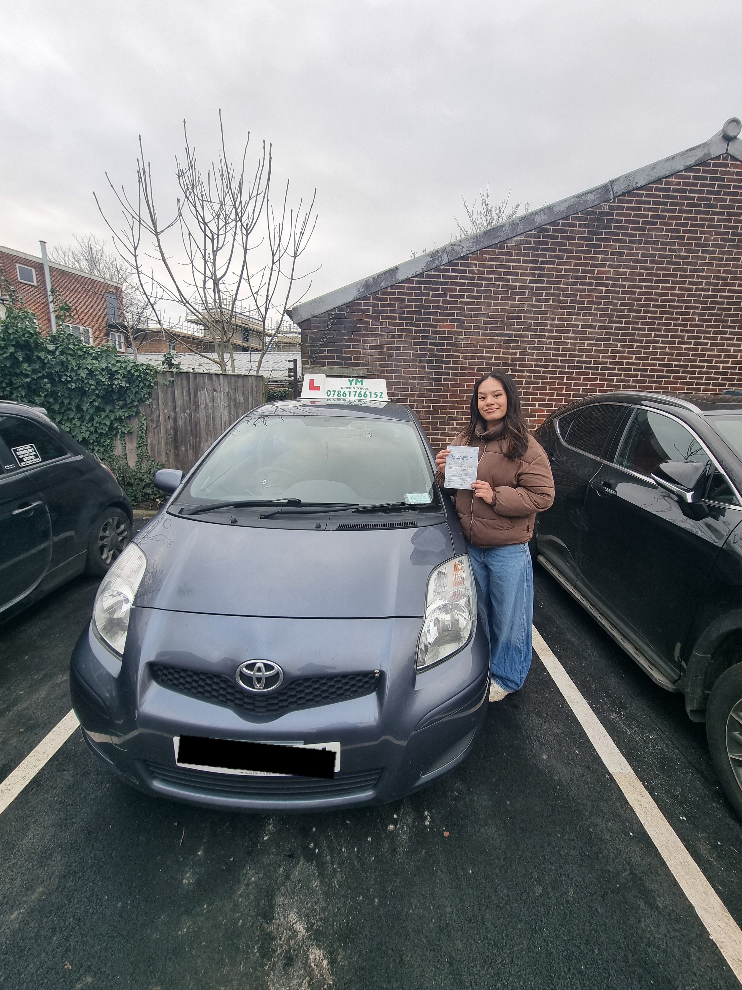 Young woman standing next to a new gray Toyota rental car with an L-plate on top, holding a paper and smiling, parked in a parking lot next to other vehicles, with a brick building and leafless tree in the background under a cloudy sky.
