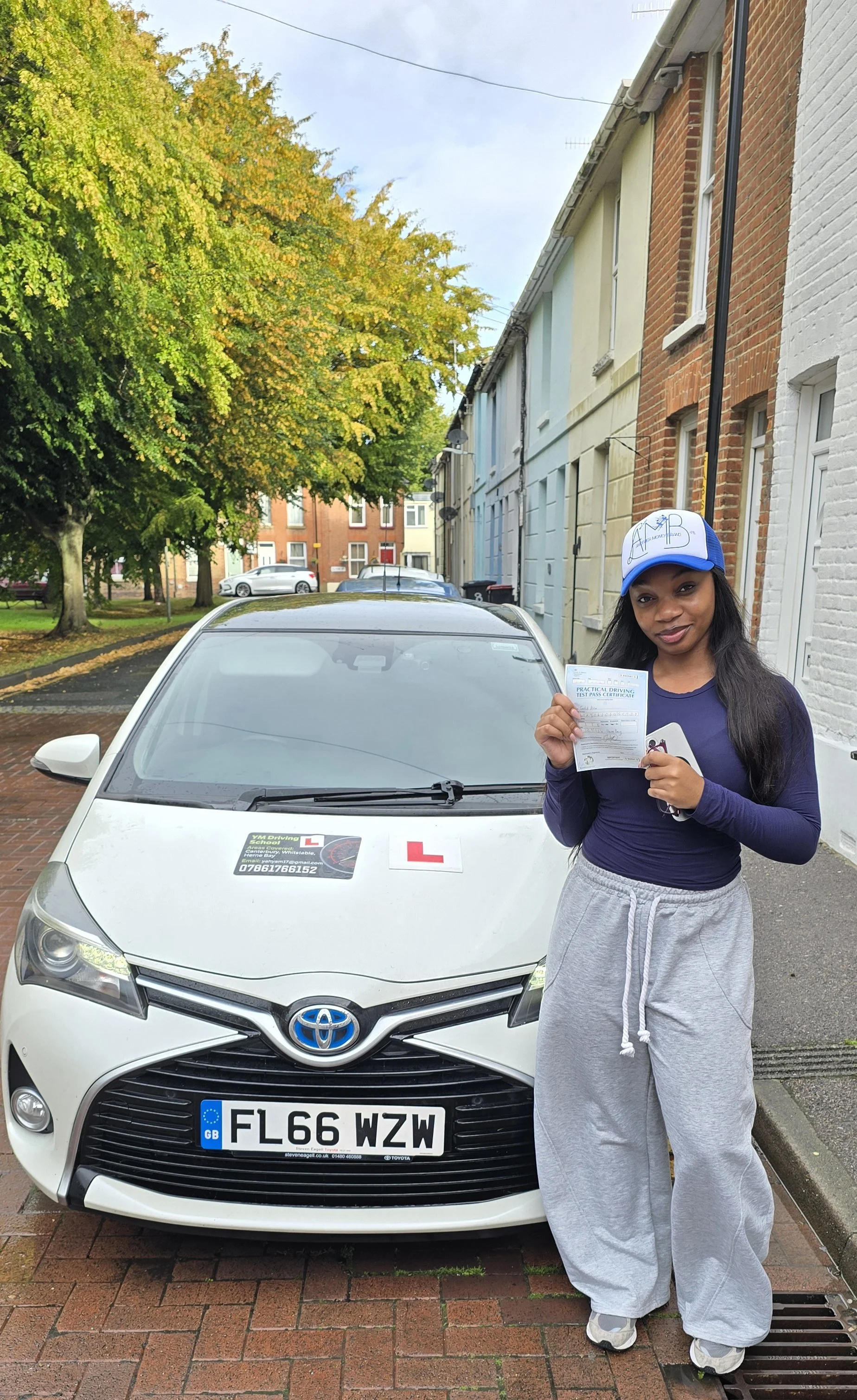 A young woman standing next to a white Toyota car, holding a practice driving test certificate. The car has "L" plates and a sticker for a driving school. She is wearing a blue and white cap, a dark long-sleeved shirt, and light gray sweatpants, smil