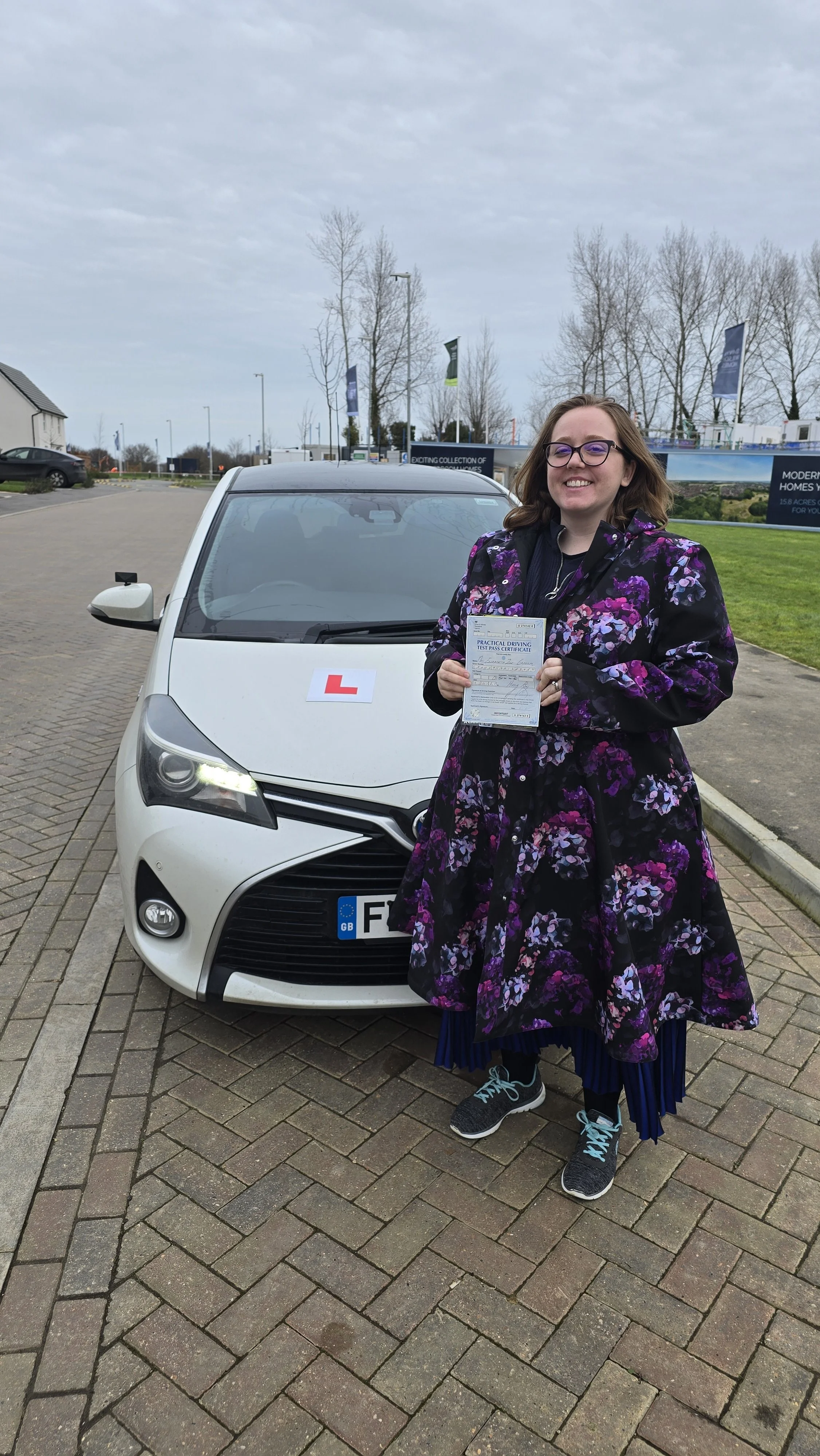 A woman holding a driving test pass certificate standing in front of a white car with an 'L' learner driver sign on the hood. She is smiling and wearing a black and purple floral coat, dark blue skirt, and sneakers. The background shows a parking lot