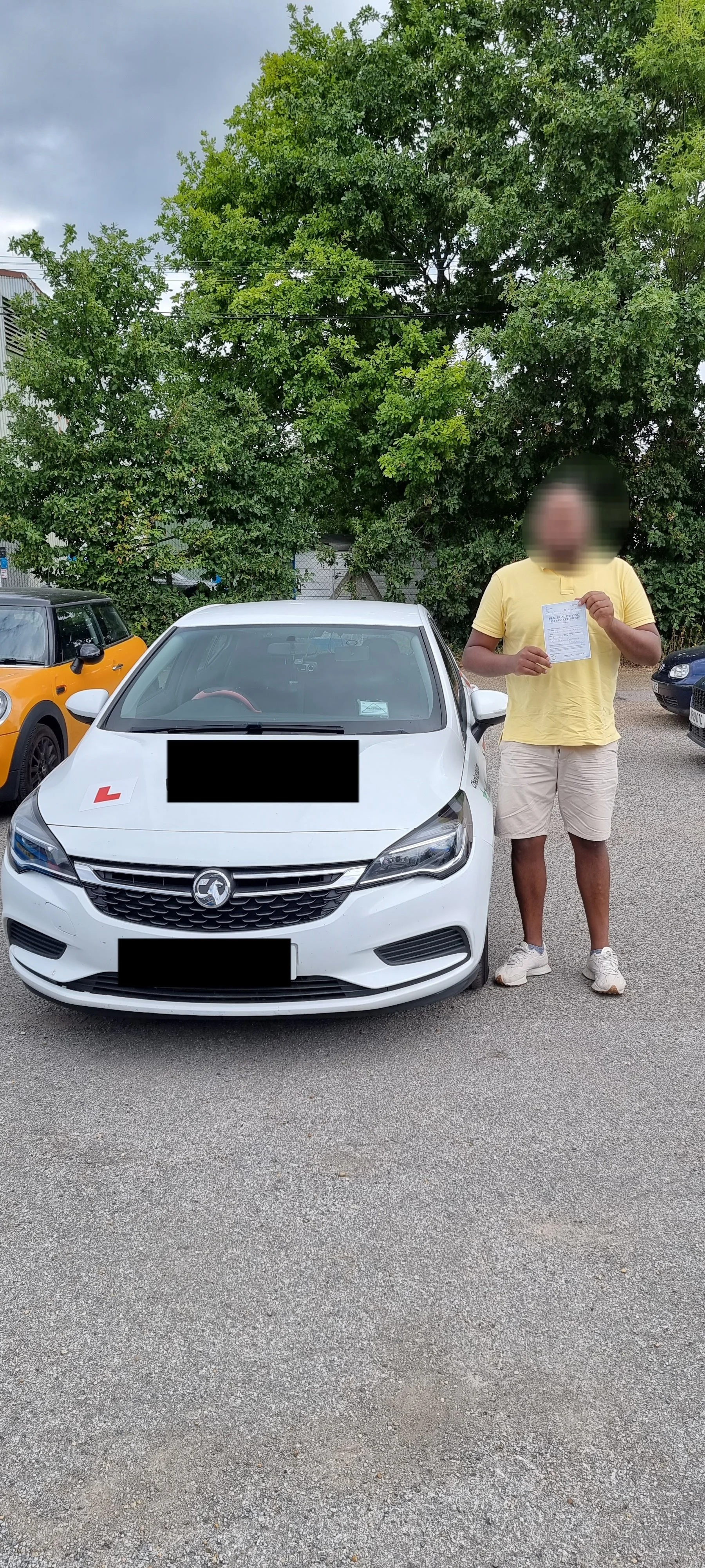 A person standing next to a white car holding a document, parked in an outdoor lot with other cars and greenery in the background.
