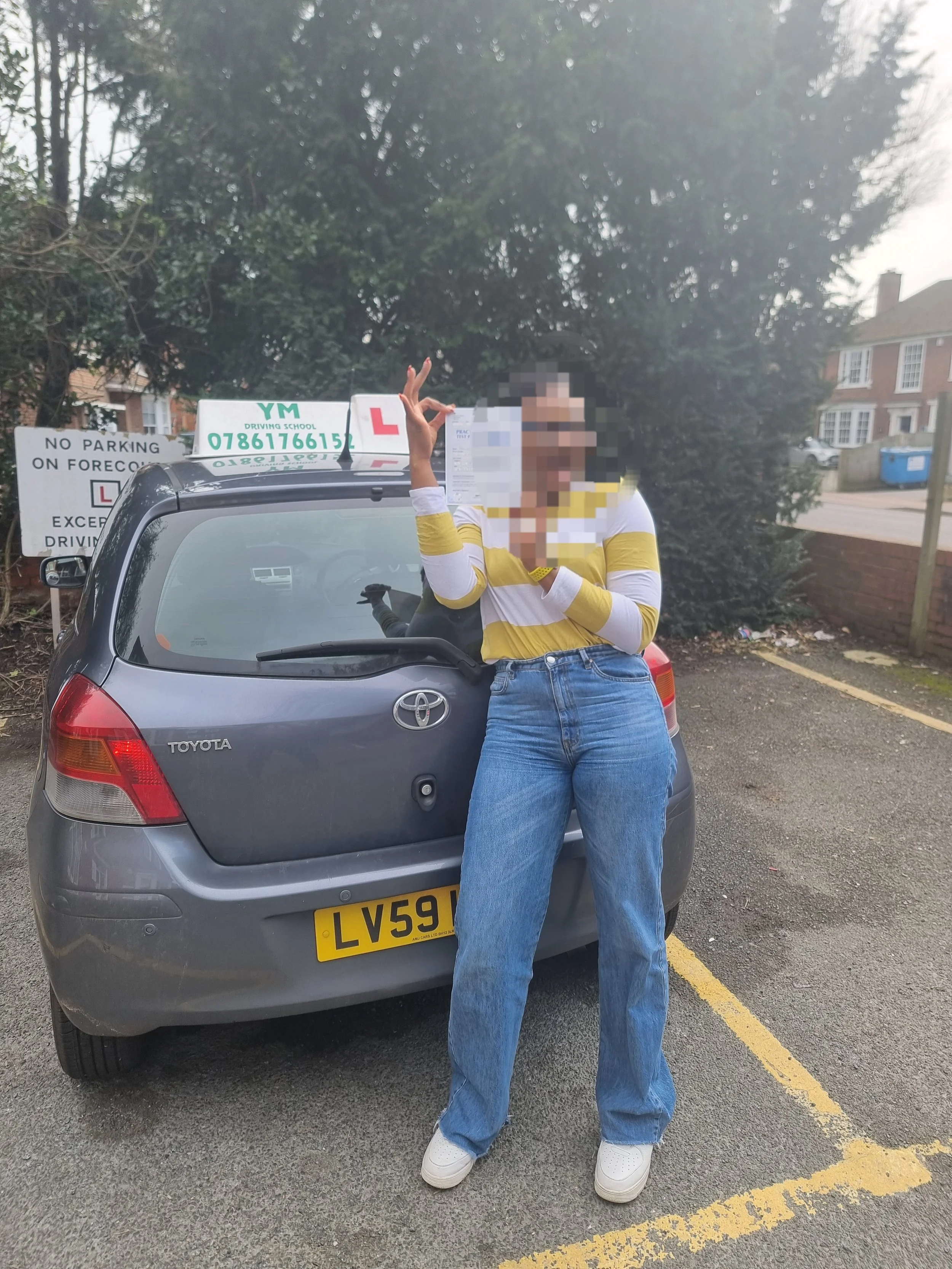 A woman standing in front of a gray Toyota car, holding a learner's driver's permit, with a driving school sign on top of the car, in a parking lot with residential houses and trees in the background.